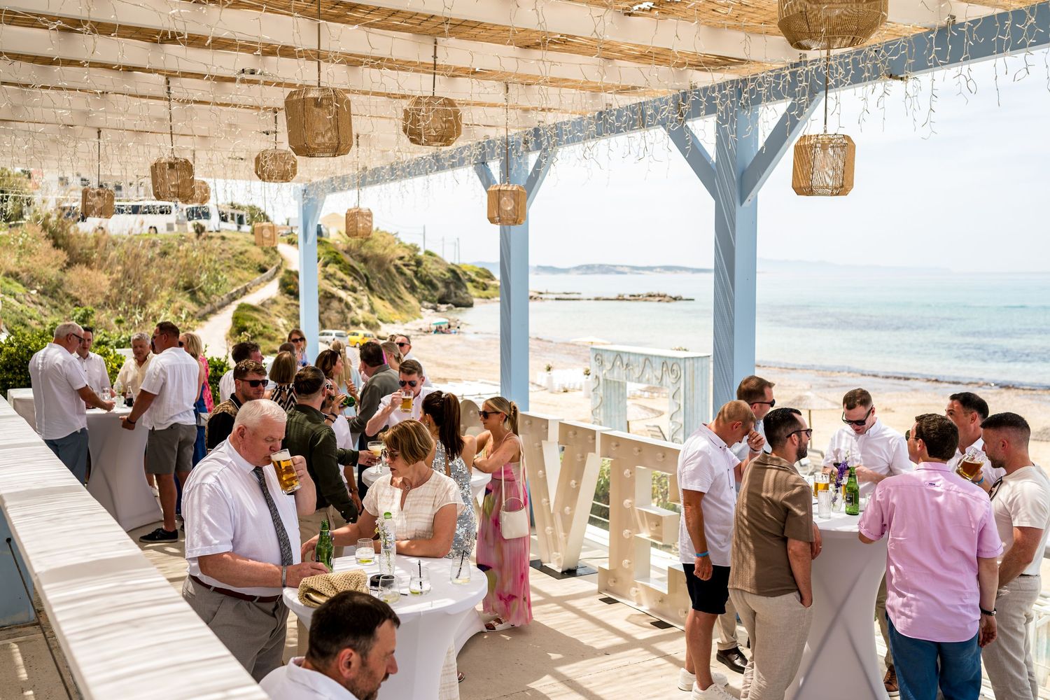 Guests mingling at an outdoor beachfront reception under wooden pergola.