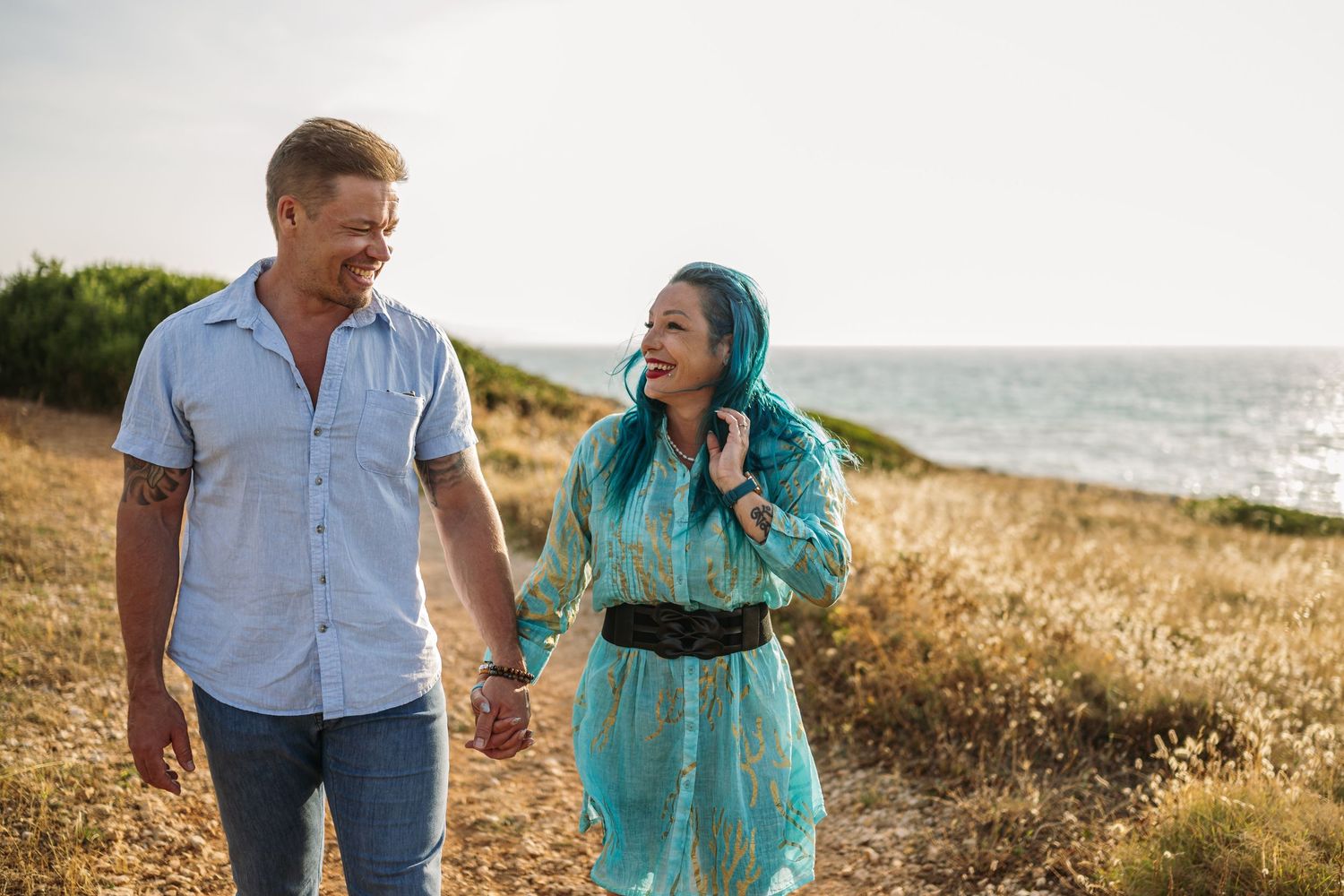 A couple takes a romantic stroll through seaside grass at sunset.