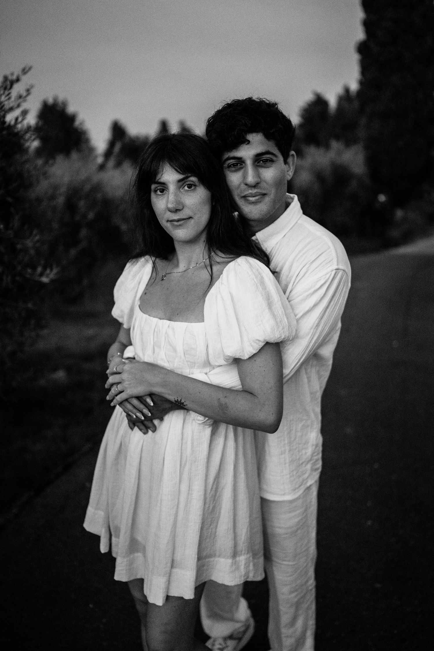 A romantic black and white portrait of a couple in white clothing embracing on a garden path.