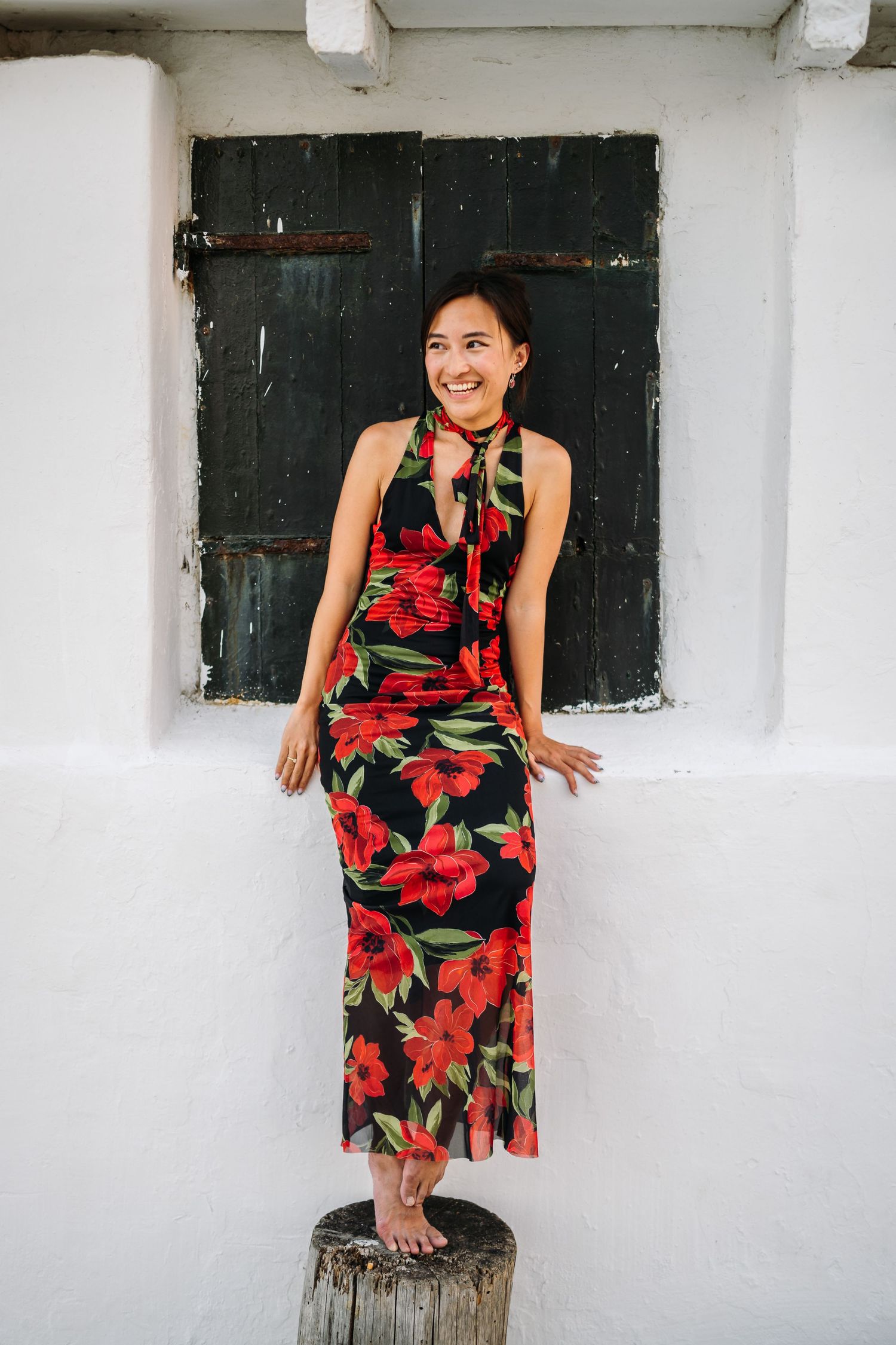 A woman in a red floral dress poses on a wooden post against a white wall with dark shutters.