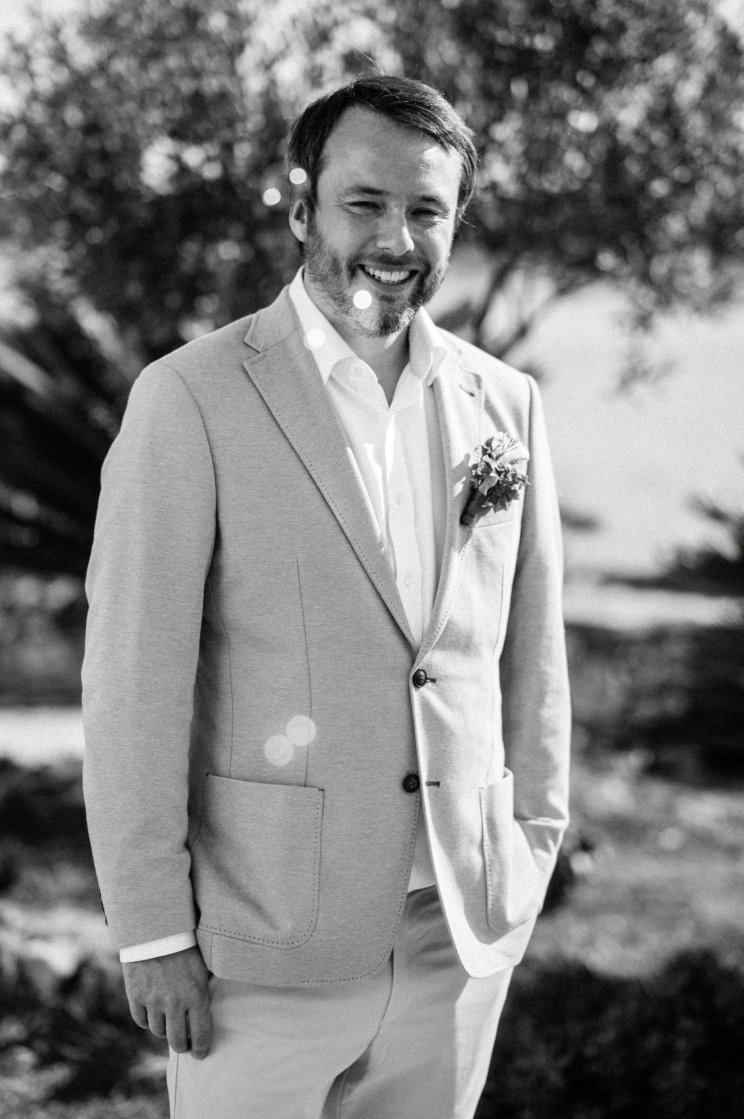 A groom in a light beige suit and white boutonniere smiles outdoors on his wedding day.