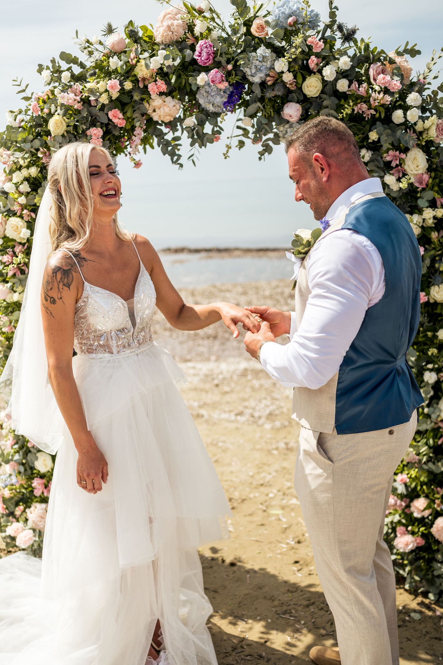 A beautiful beach wedding ceremony under a floral arch with pink and white roses and greenery.