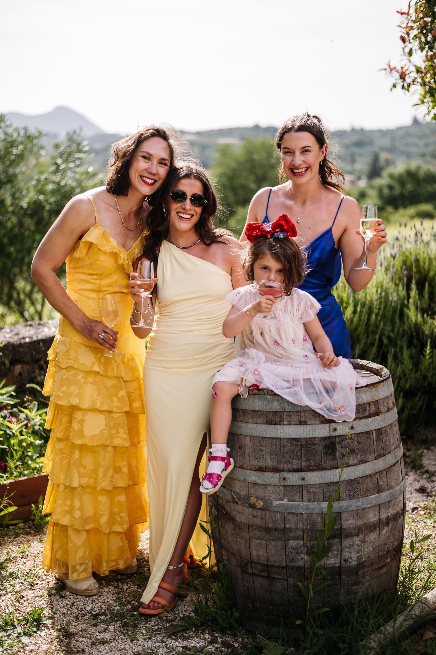 Bridesmaids in yellow and cream dresses pose with flower girl by wooden barrel.