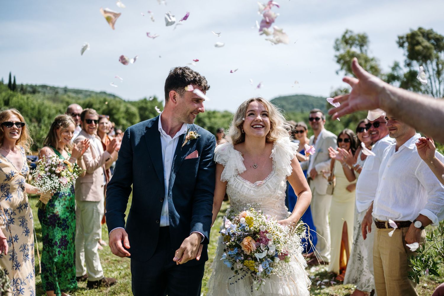 Wedding party celebrates with confetti against a backdrop of rolling hills and blue sky.