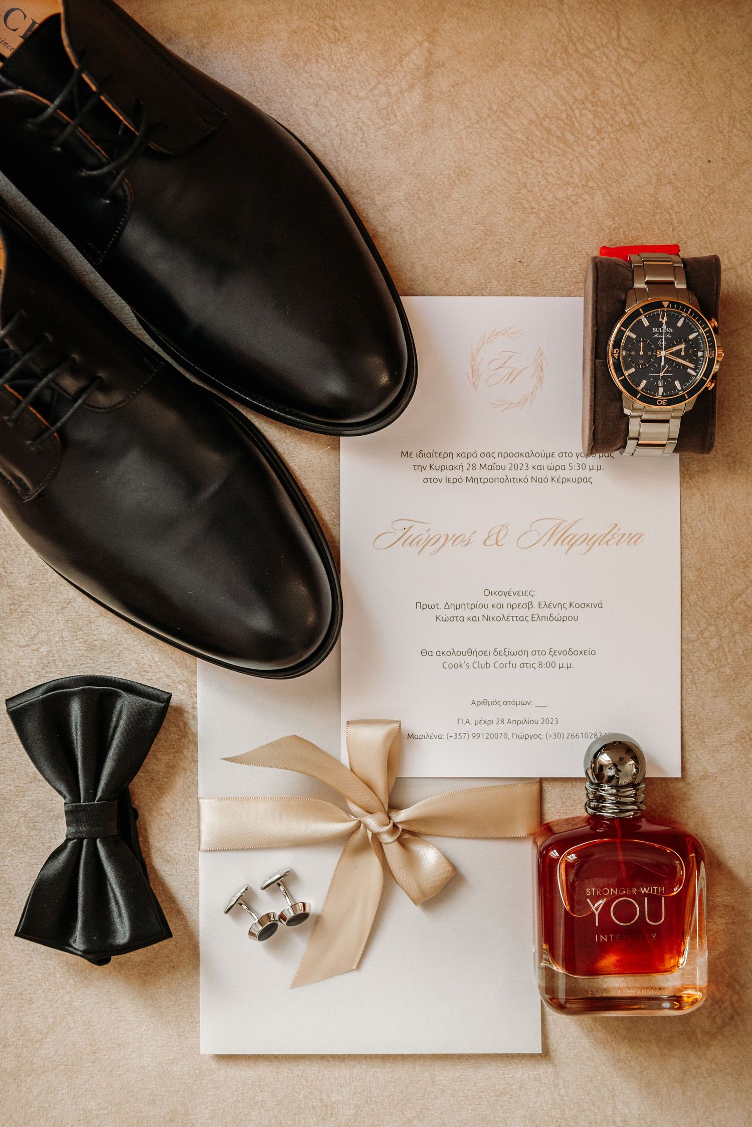 Wedding accessories including dress shoes, bow tie, watch, invitation and perfume arranged on beige surface.
