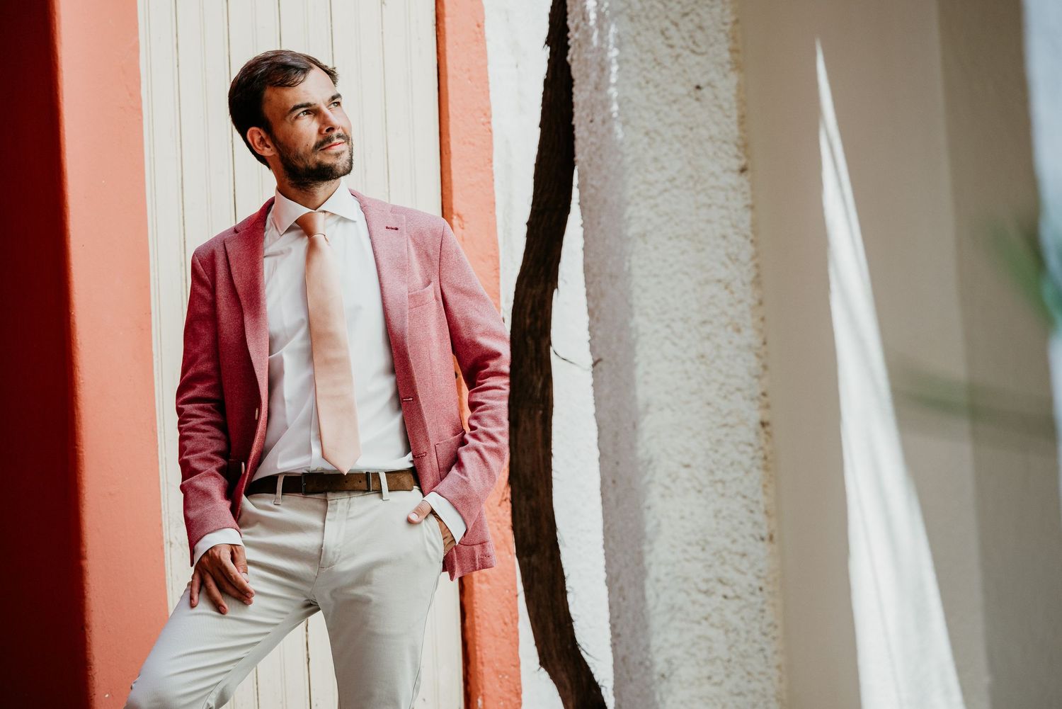 A person in a burgundy suit jacket and white shirt poses against a light colored wall.