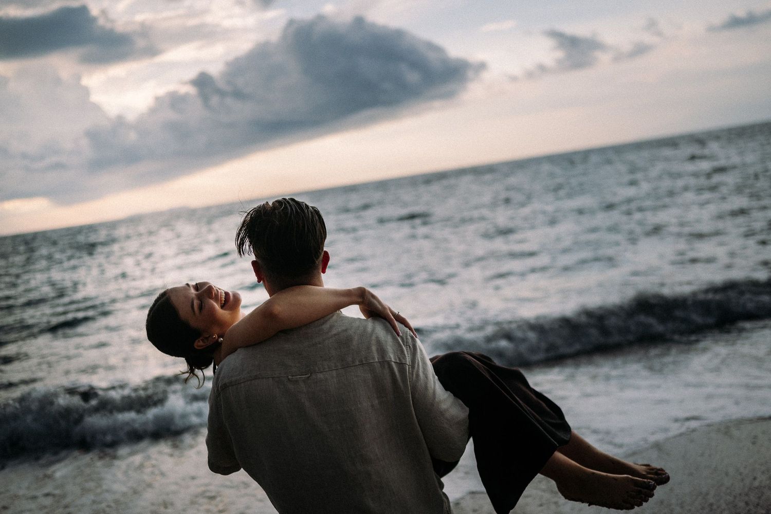 Romantic couple embracing on a beach at sunset, man carrying woman in his arms near ocean waves.