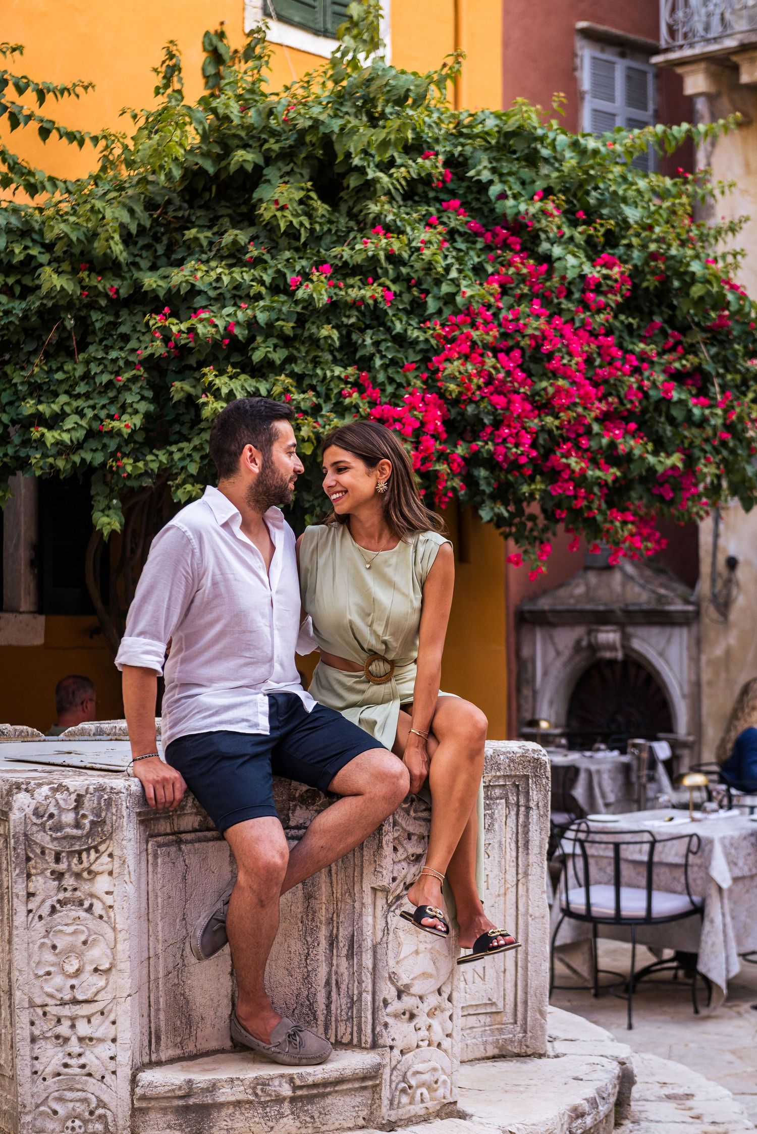 Couple sitting on stone wall against orange building with blooming bougainvillea.