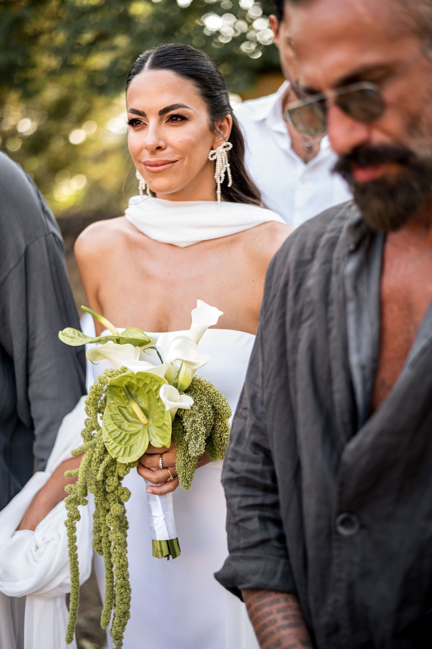 A bride in white holds a bouquet with striking green ribbons during an outdoor ceremony.