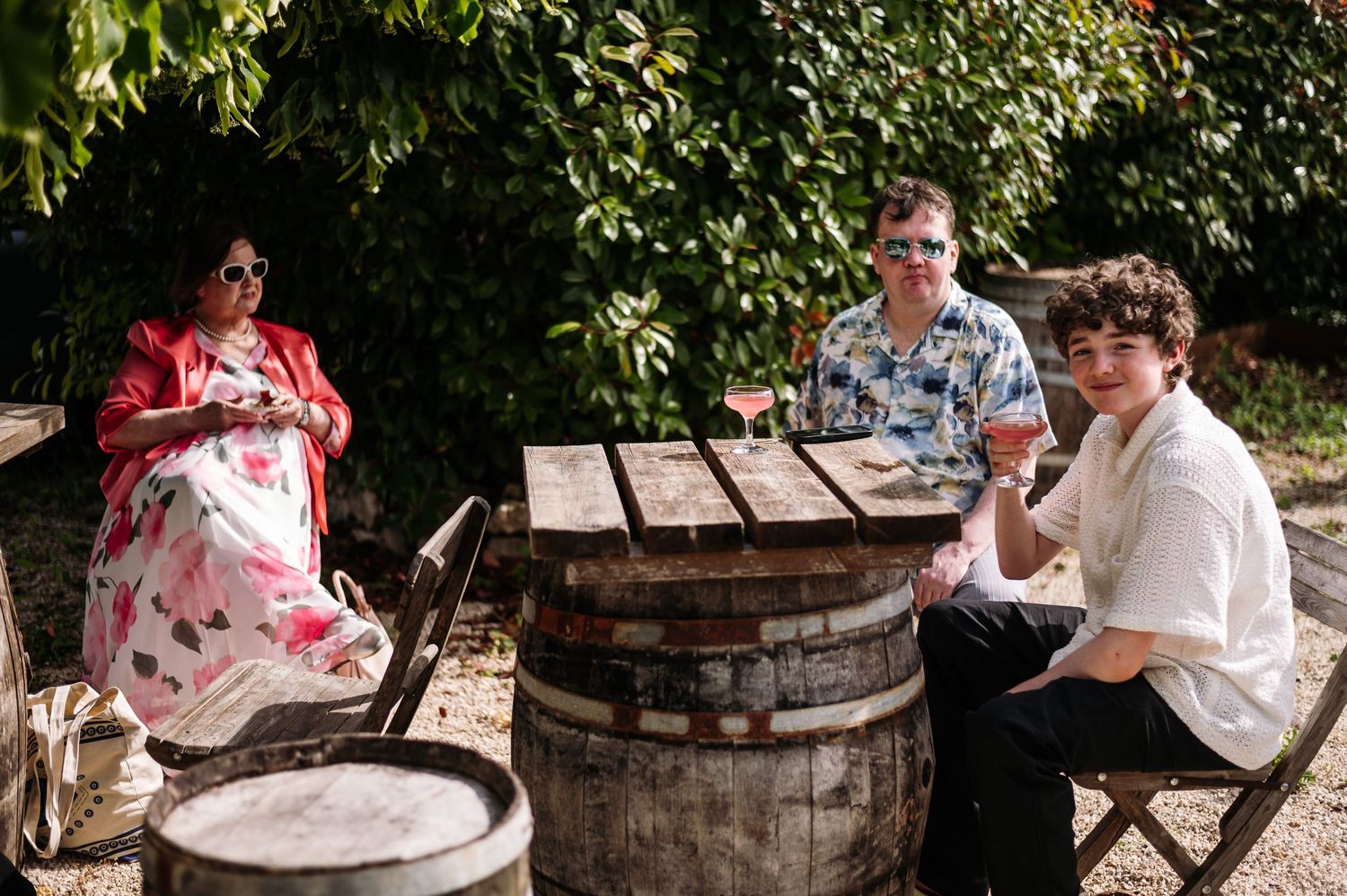 Wedding party gathers around rustic wine barrels in an outdoor vineyard setting.