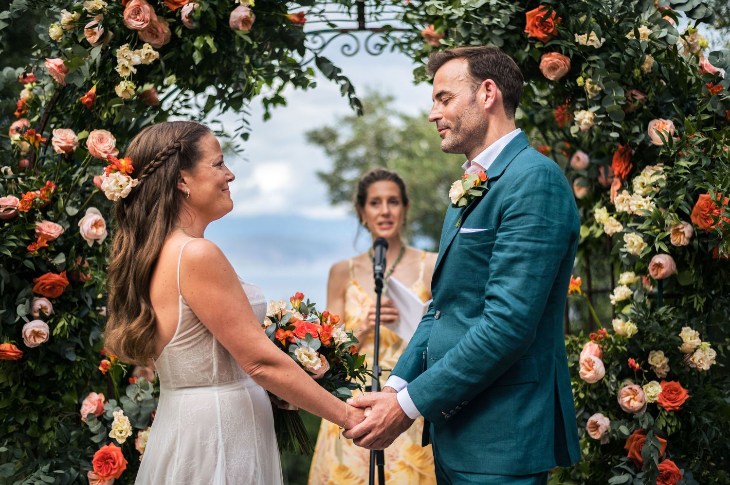 Couple exchanging vows under a floral arch with coral roses in a garden setting.