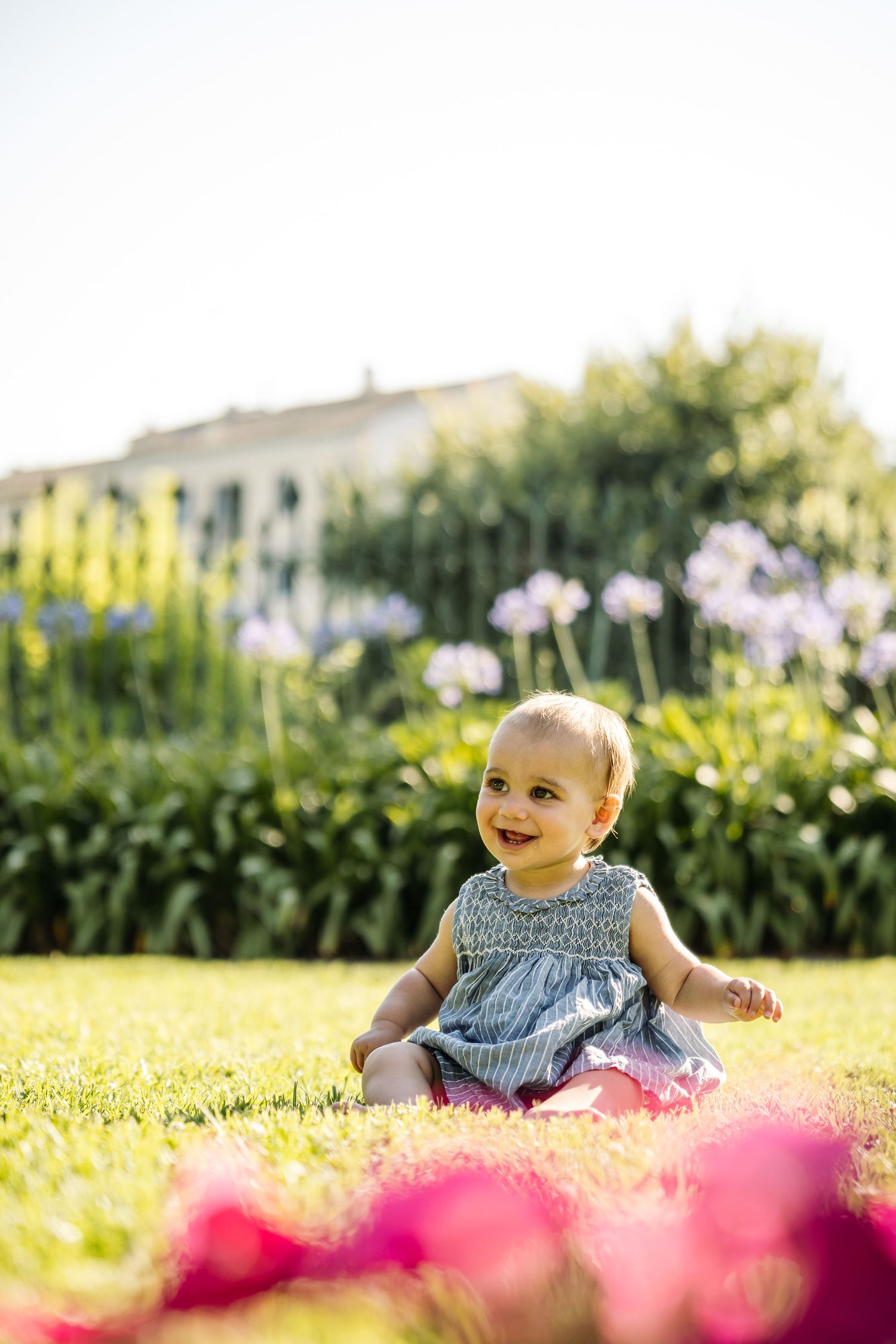 Baby in blue dress sitting on grass with blooming flowers in garden setting.