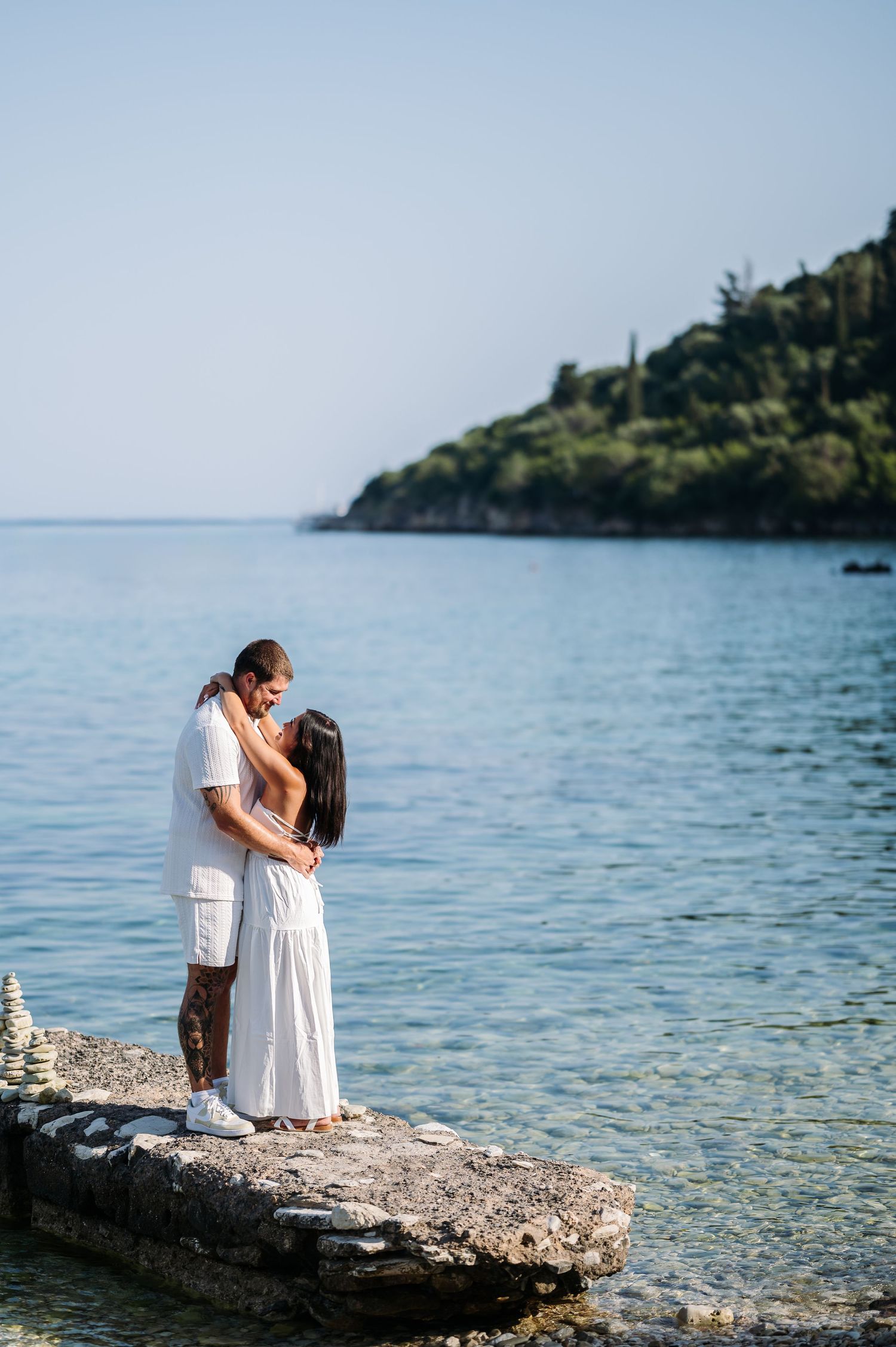A romantic couple in white attire embracing on a rocky shoreline with blue waters in the background.