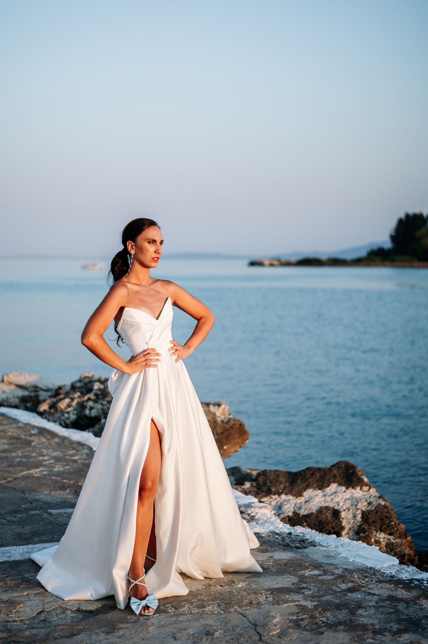 Elegant white wedding dress photographed against a scenic rocky coastline and blue ocean backdrop.