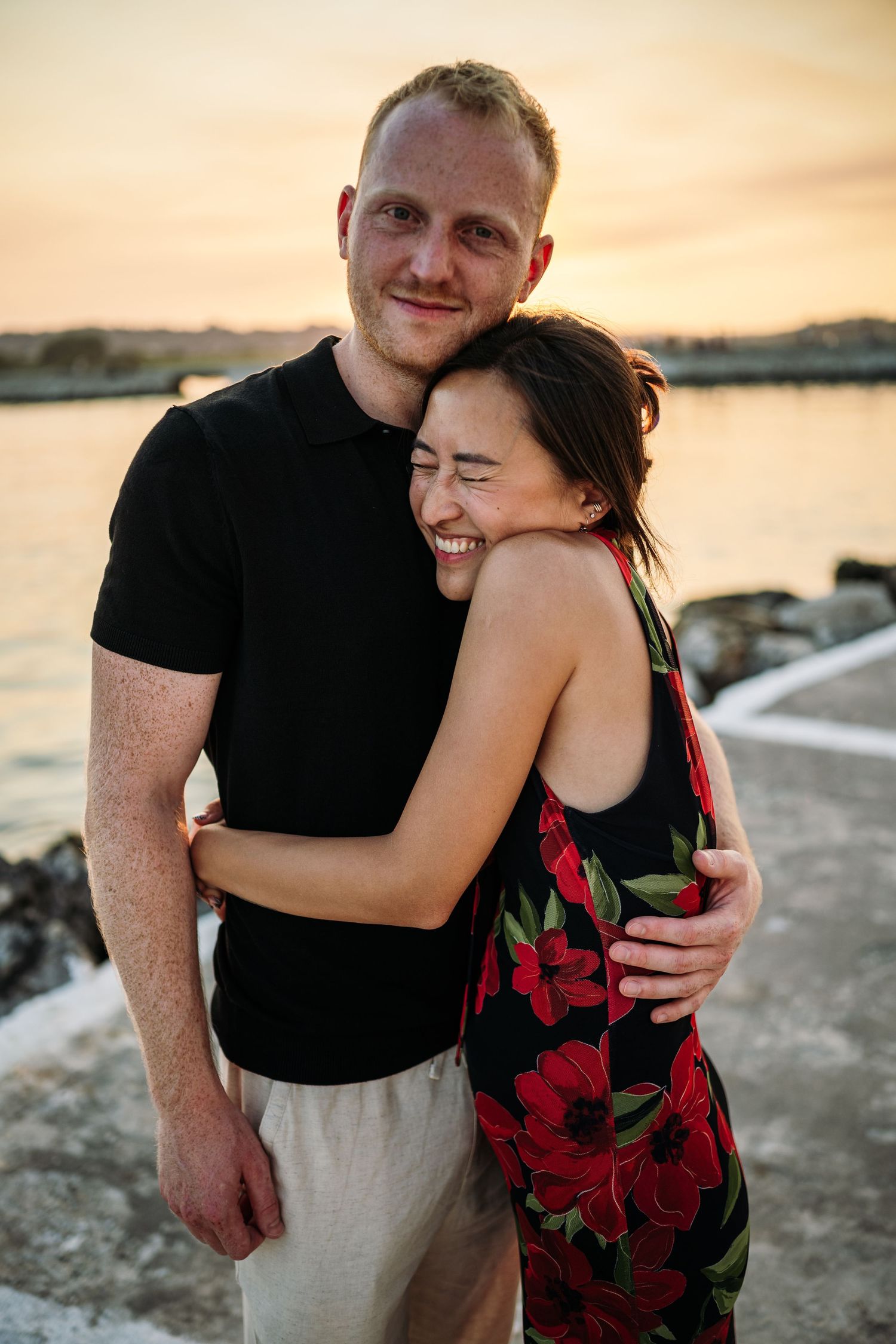 A couple embracing at sunset by the ocean wearing black and floral dress.