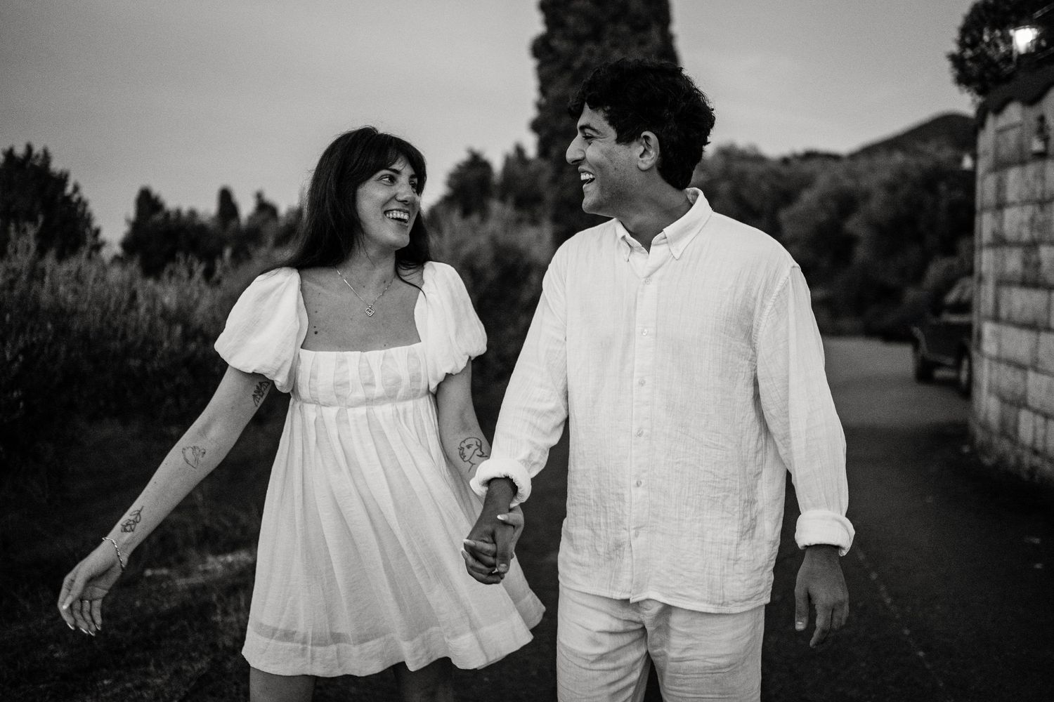 A playful black and white photo of a couple in white outfits walking and laughing together on a stone path.