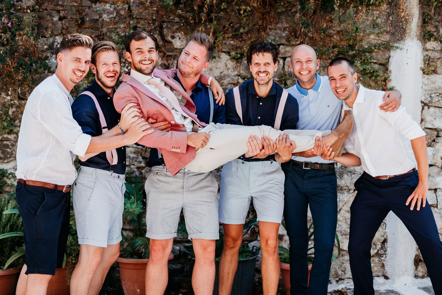 Group of wedding guests posing and laughing together against stone wall.