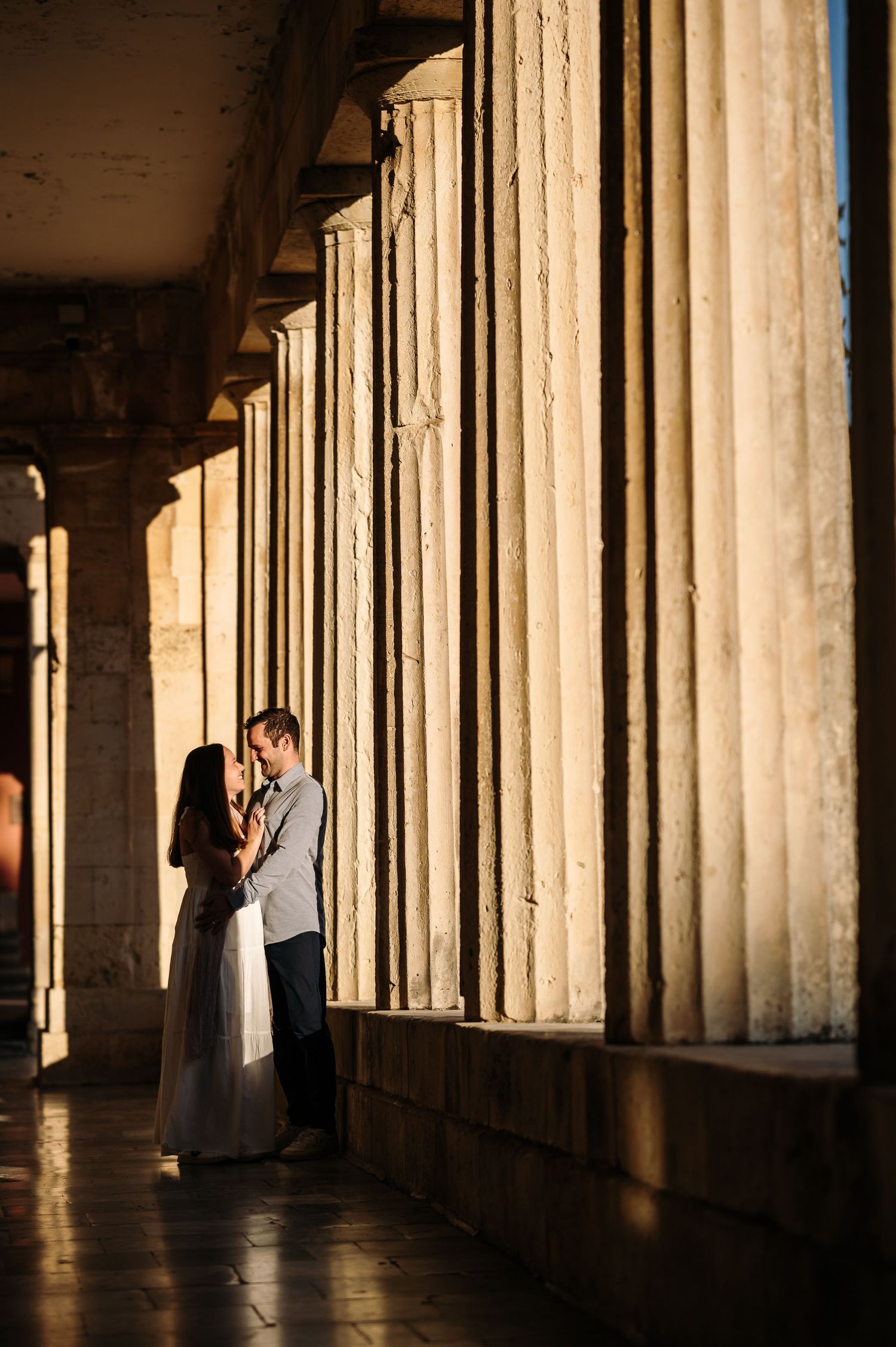 A couple embraces between dramatic stone columns with sunlight creating striking shadows.