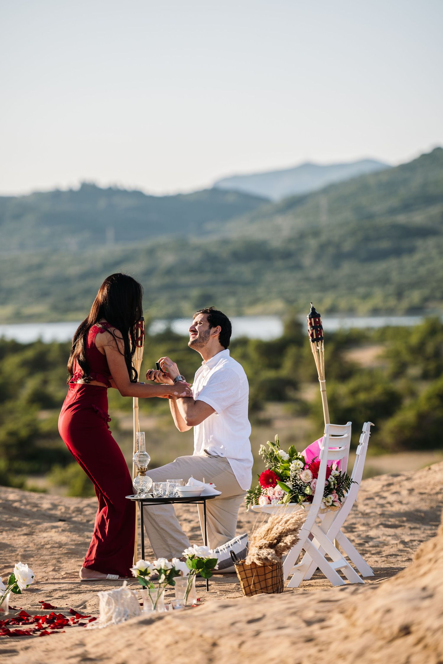 A romantic sunset proposal setup on a beach with candles, flowers and mountains in the background.