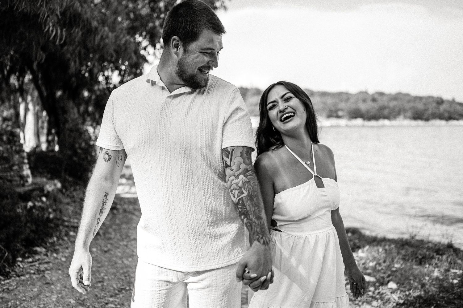 A black and white photo of a couple laughing together while walking along a waterfront path.
