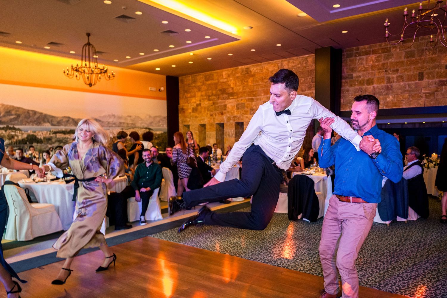 Wedding guests performing energetic dance moves on illuminated reception dance floor.