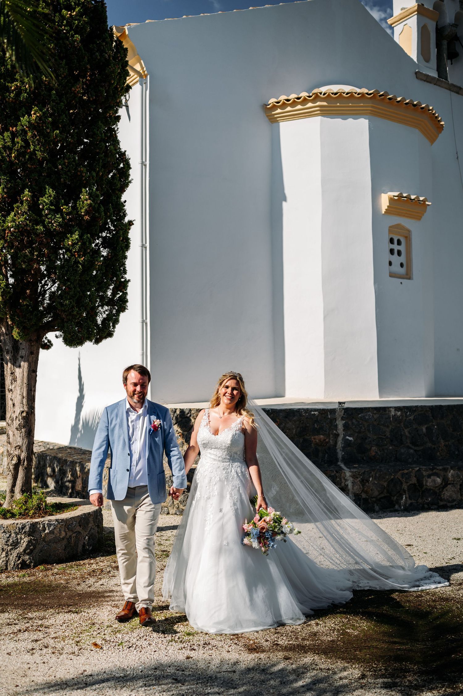 Wedding couple walking along white Mediterranean chapel exterior.