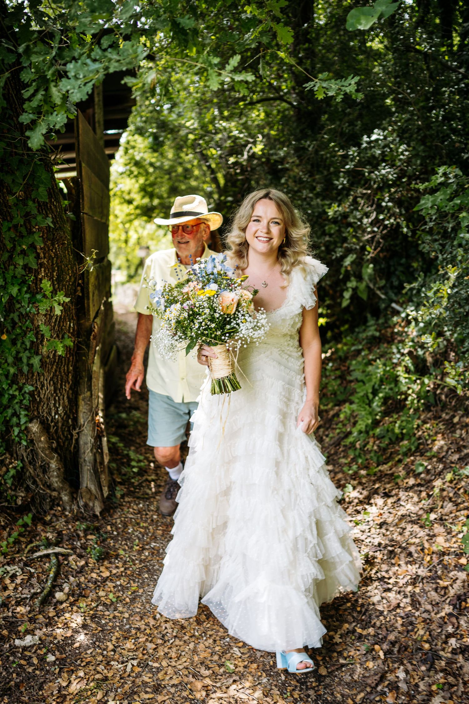 Sunlit wooded path with figures walking through dappled shade carrying floral bouquets.