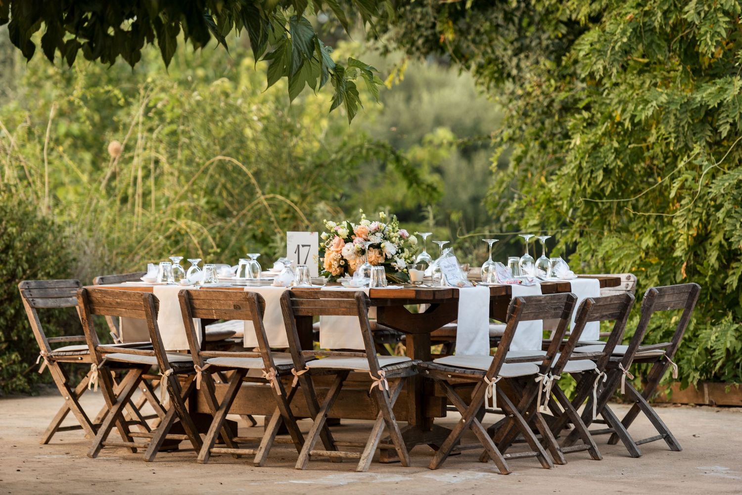 Rustic outdoor wedding reception table setup with wooden chairs and floral arrangements.