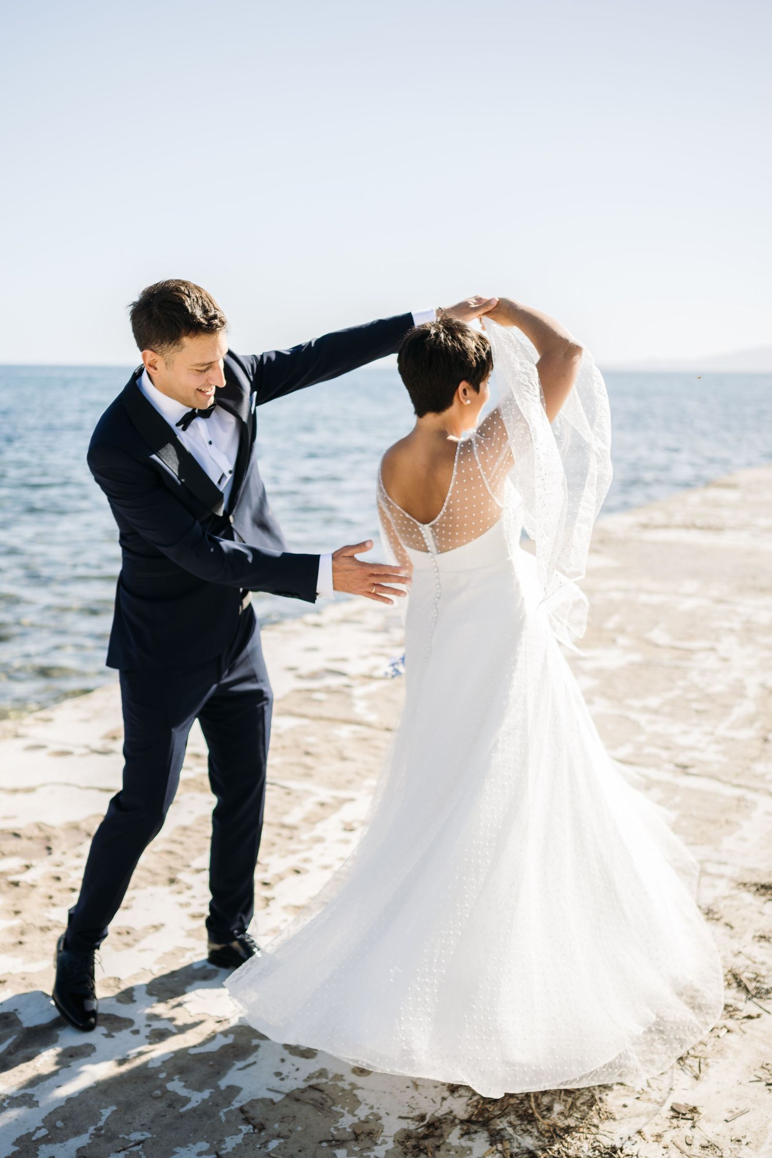 A playful wedding couple dances on a sandy beach with ocean views.