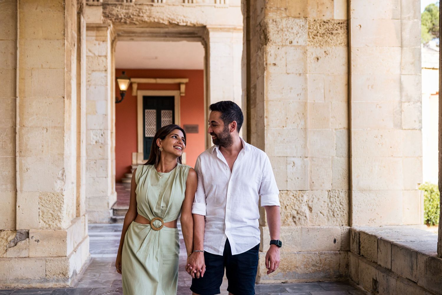 A couple walks through a historic stone colonnade with warm sunlight filtering through.