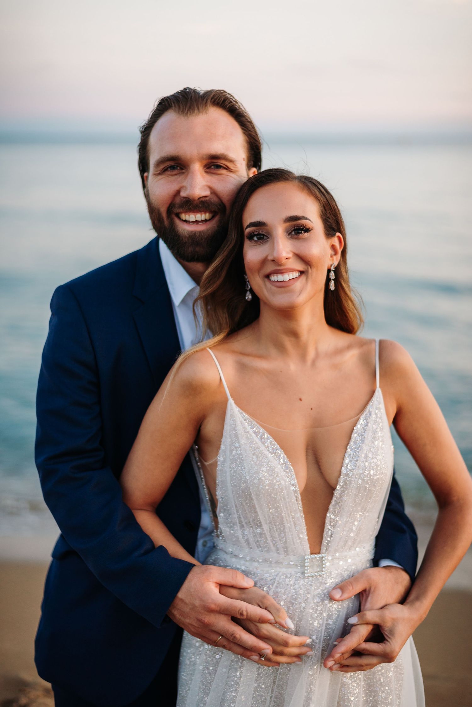 A romantic beach photoshoot featuring a couple in wedding attire embracing by the ocean at sunset.