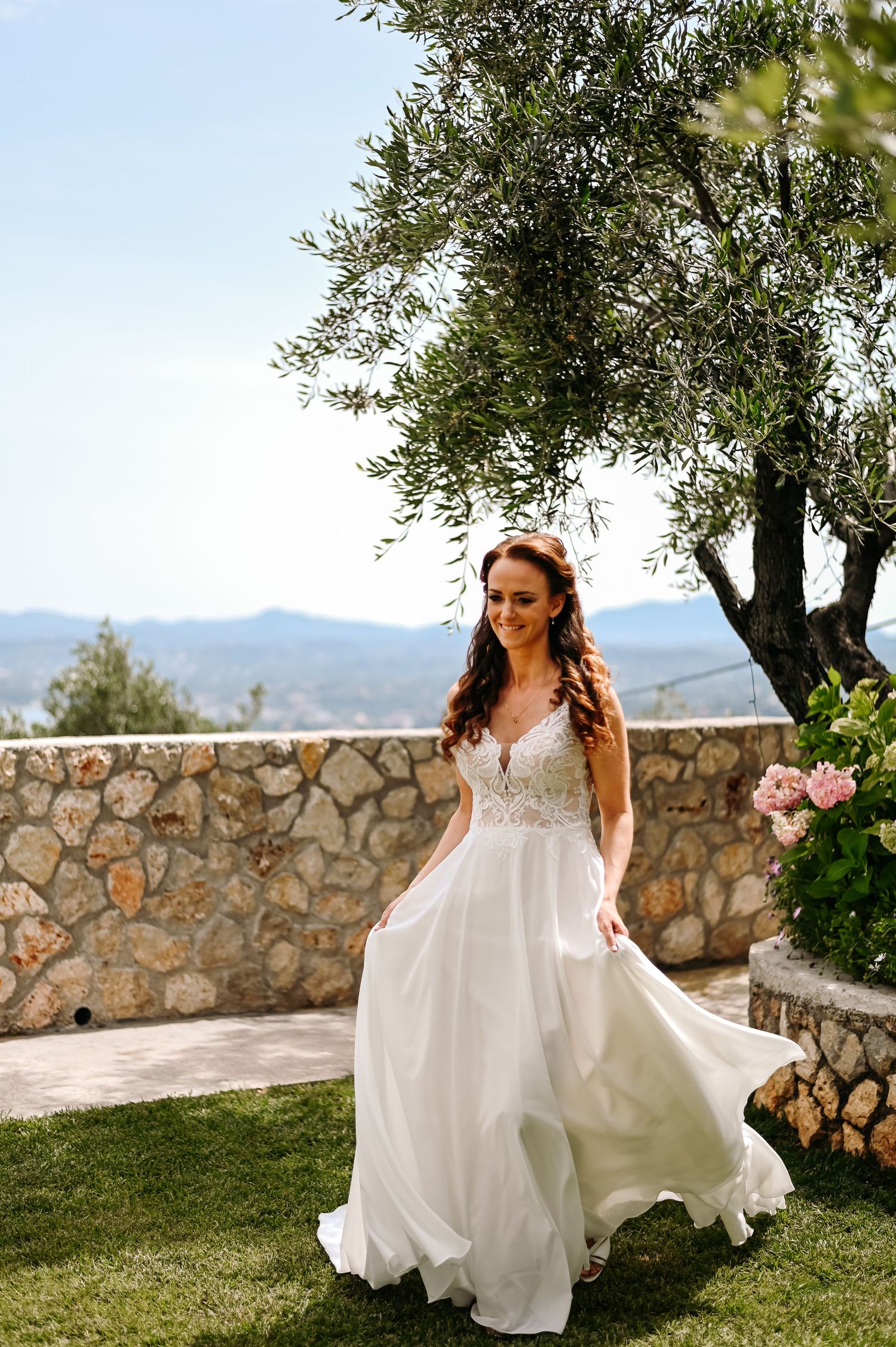A white wedding dress flows in the breeze against a mountainous backdrop and stone wall.