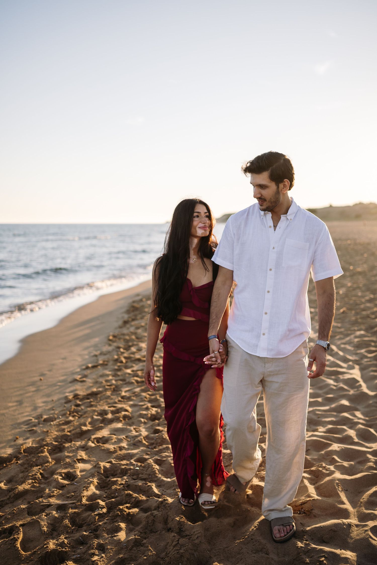 Couple in red and white outfits walking together on a sandy beach at sunset.