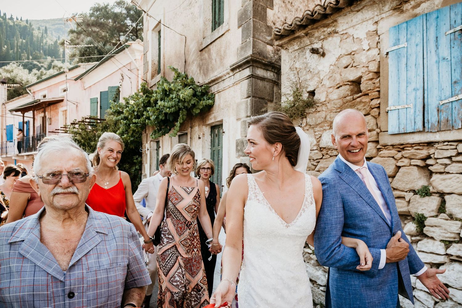 Wedding guests gather on a charming Mediterranean street with stone walls and blue shutters.