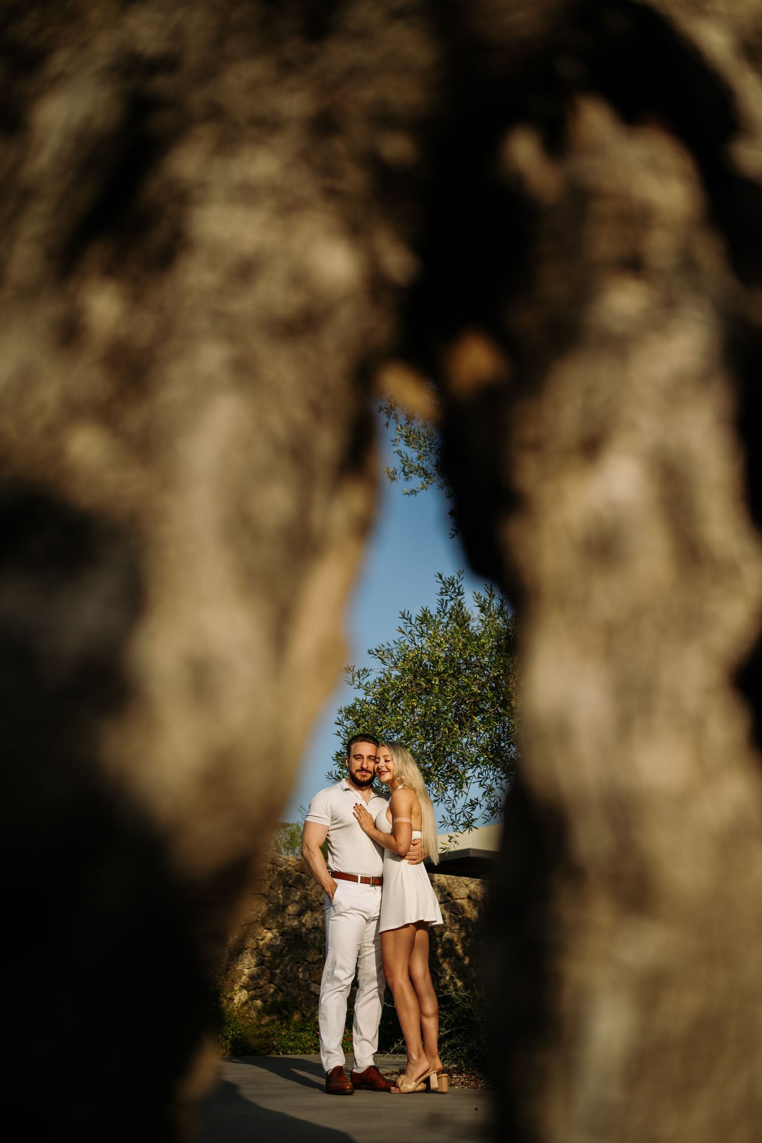A couple embraces in white outfits framed by blurred rocky foreground elements.