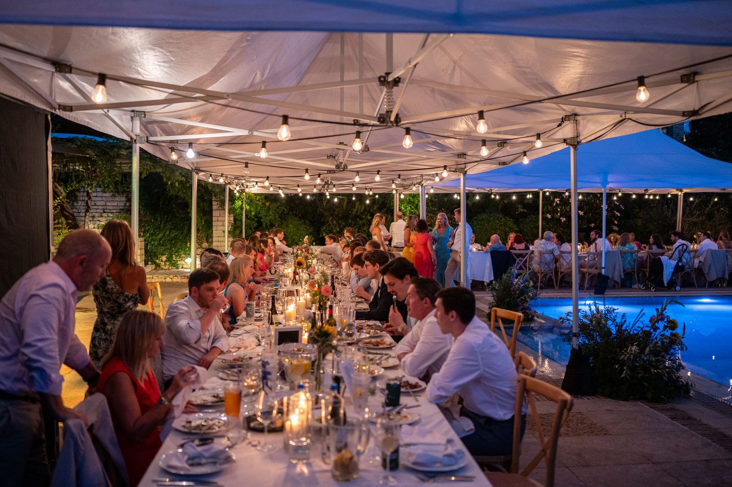 Elegant outdoor wedding reception under white tents beside illuminated swimming pool at dusk.