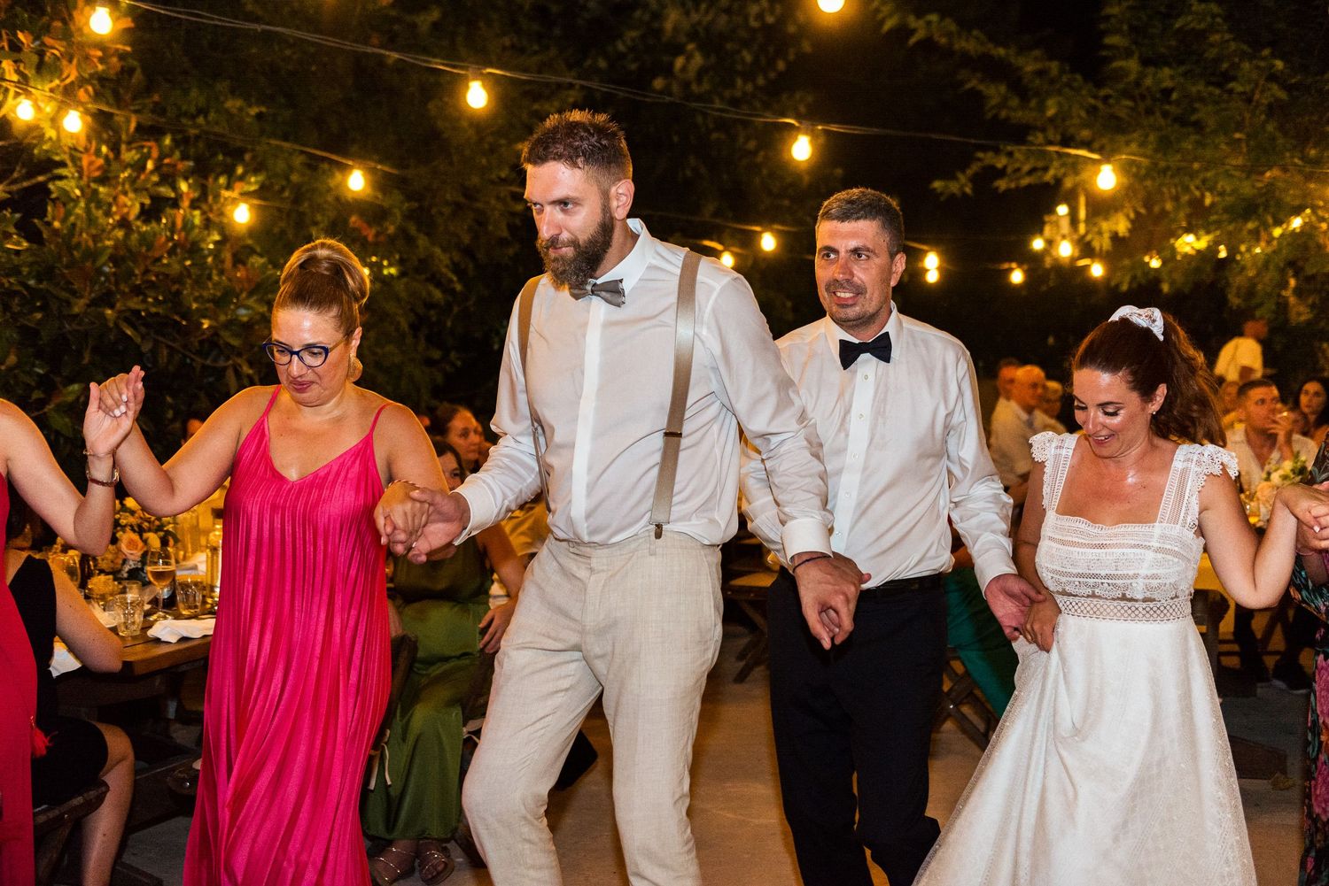 Wedding party dancing and celebrating under warm string lights at an outdoor evening reception.