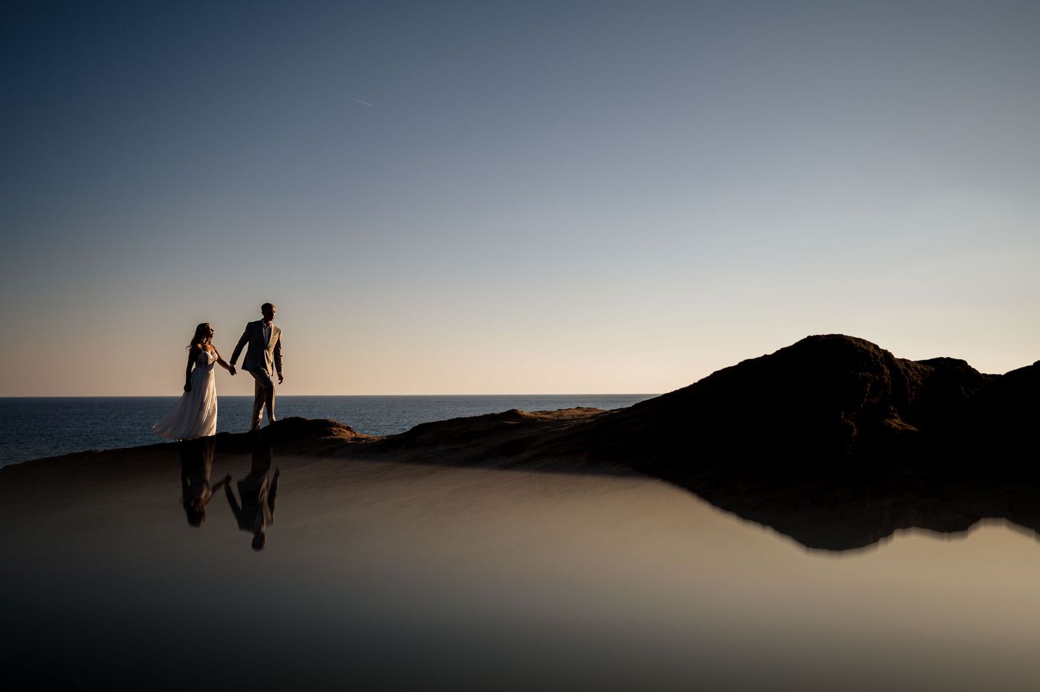 A silhouetted wedding couple stands reflected in still water at sunset by the ocean.