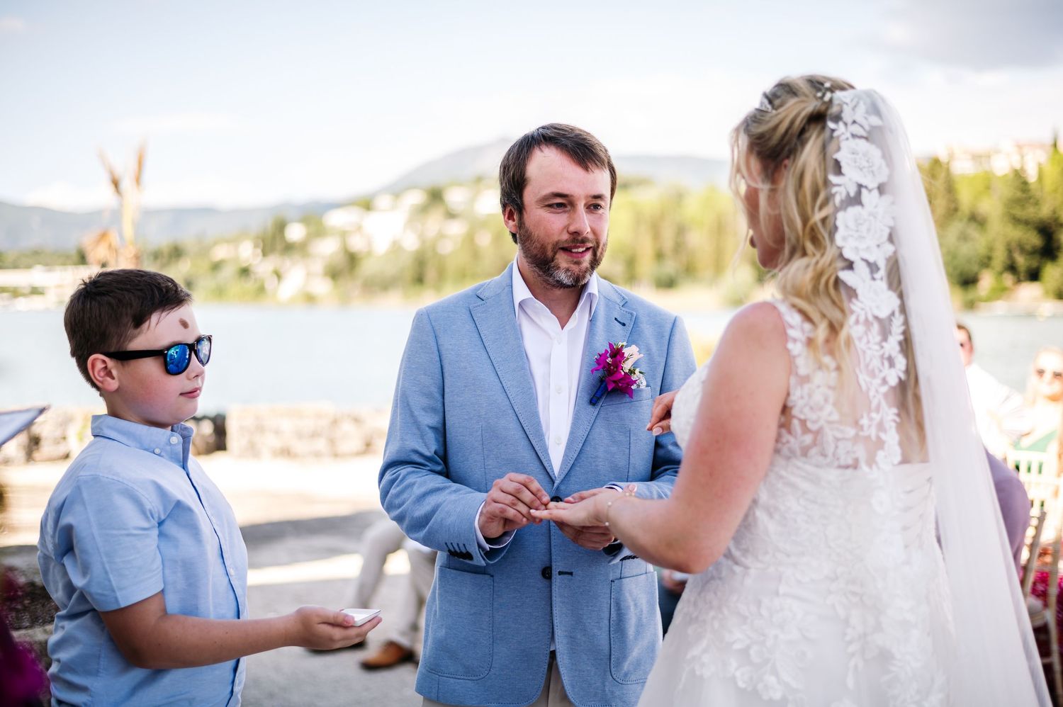 An emotional wedding ceremony moment captured in bright sunlight with pink flowers framing the scene.