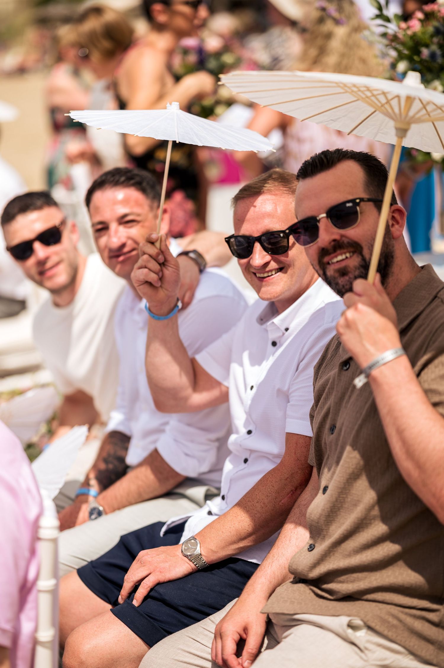 Wedding guests sit in white chairs with parasols shading them from the sun.