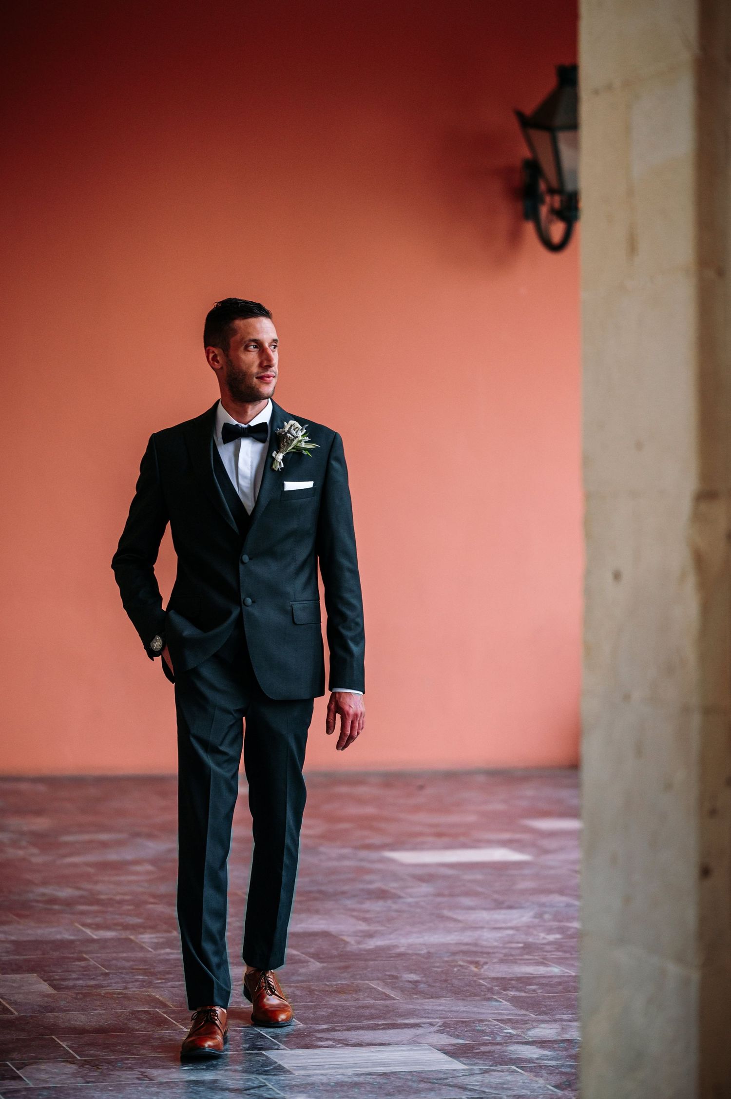 Elegant groom in classic black tuxedo poses against coral wall with vintage lantern.