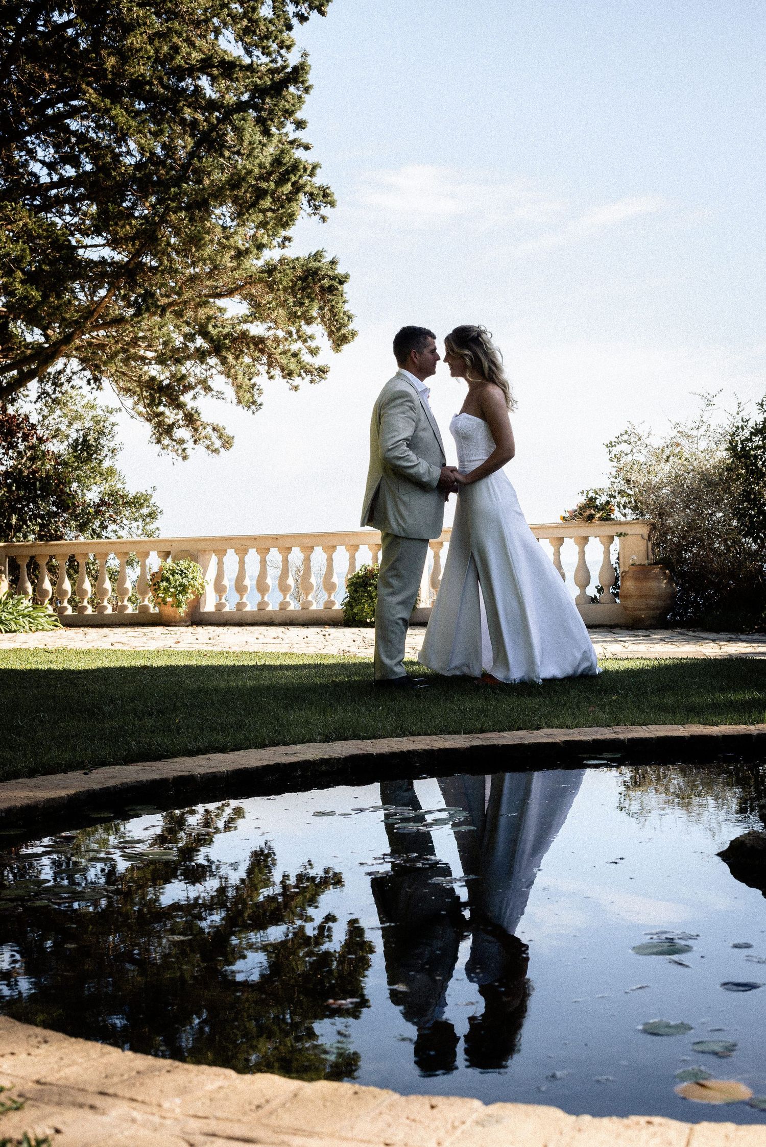 Newlyweds pose together by a reflecting pool with ornate stone balustrades at sunset.