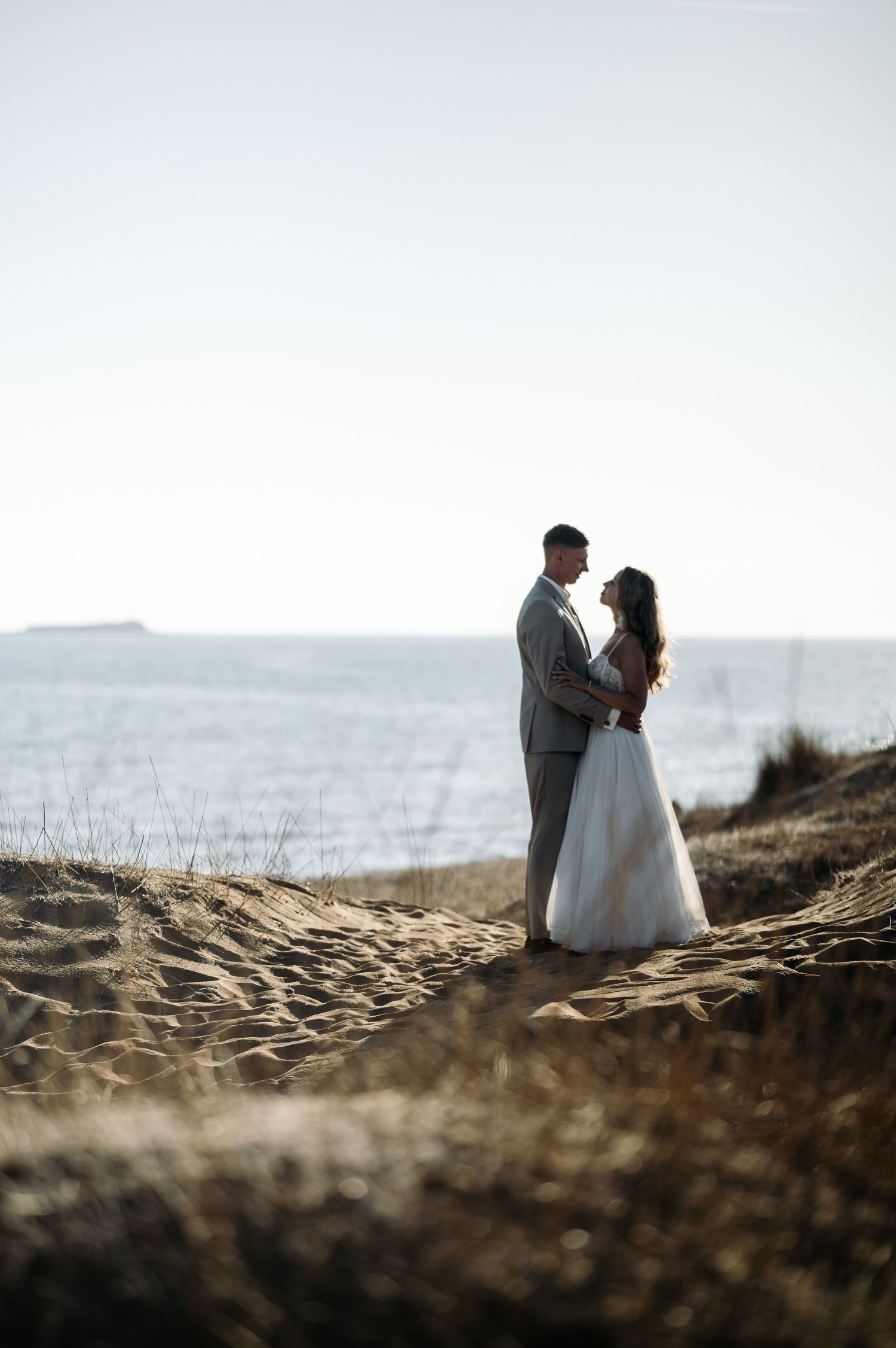 A wedding couple stands together on a sandy cliff overlooking the ocean at sunset.