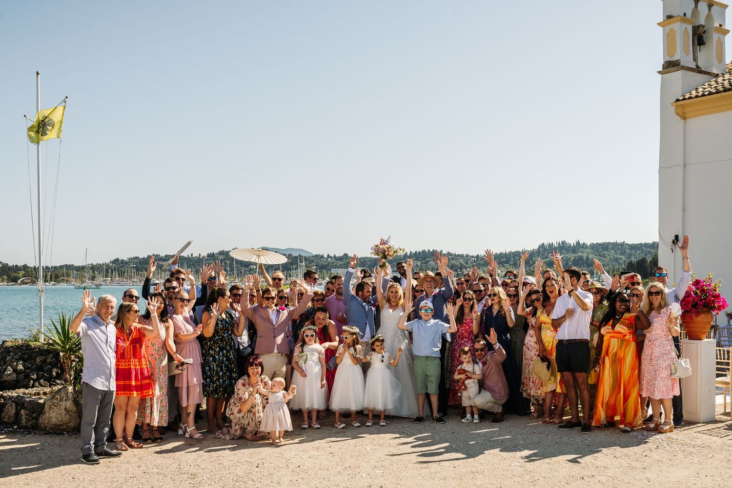 Large wedding party gathered outside a Mediterranean chapel with ocean views.