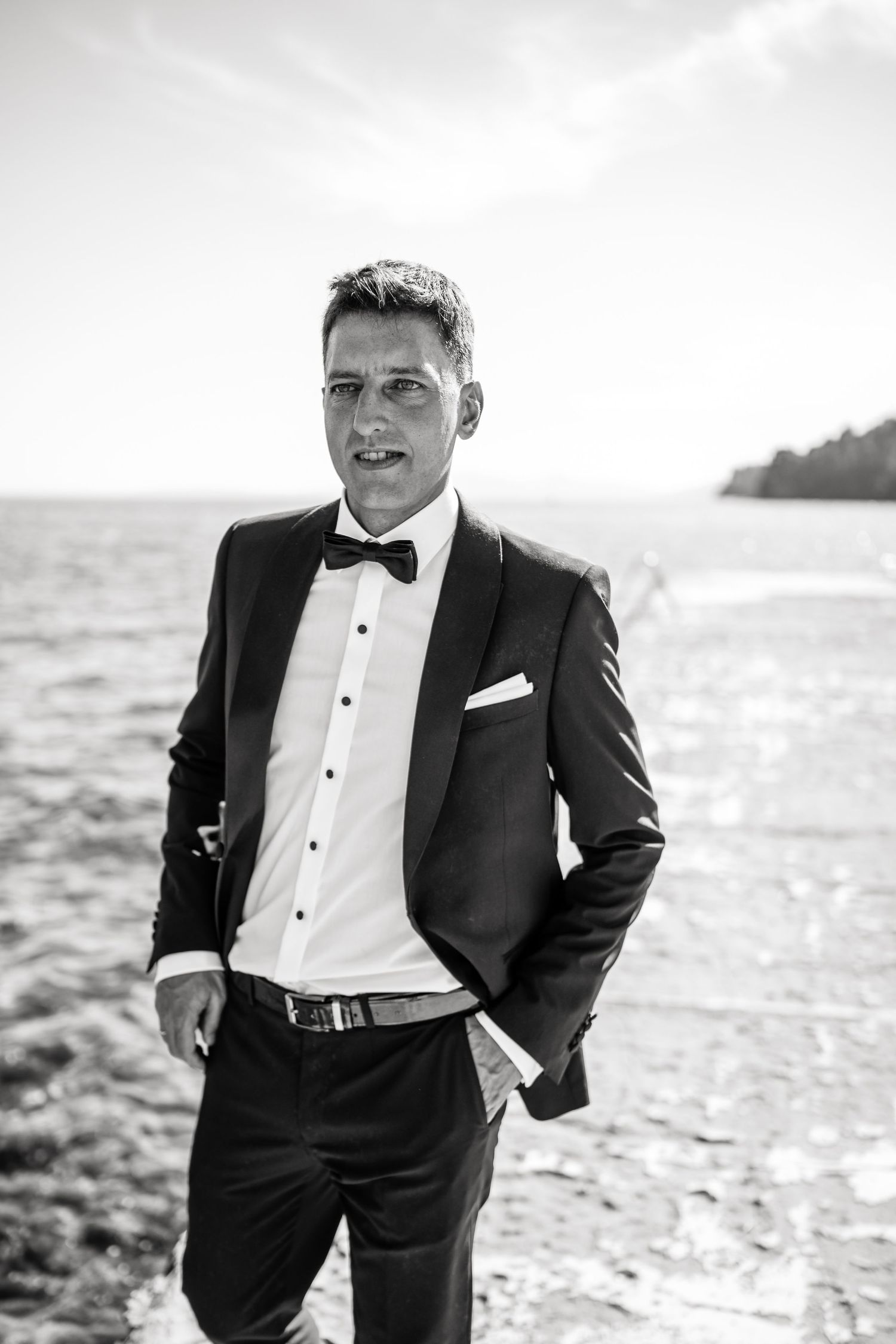 A groom in classic black tuxedo poses confidently by the ocean on a bright day.
