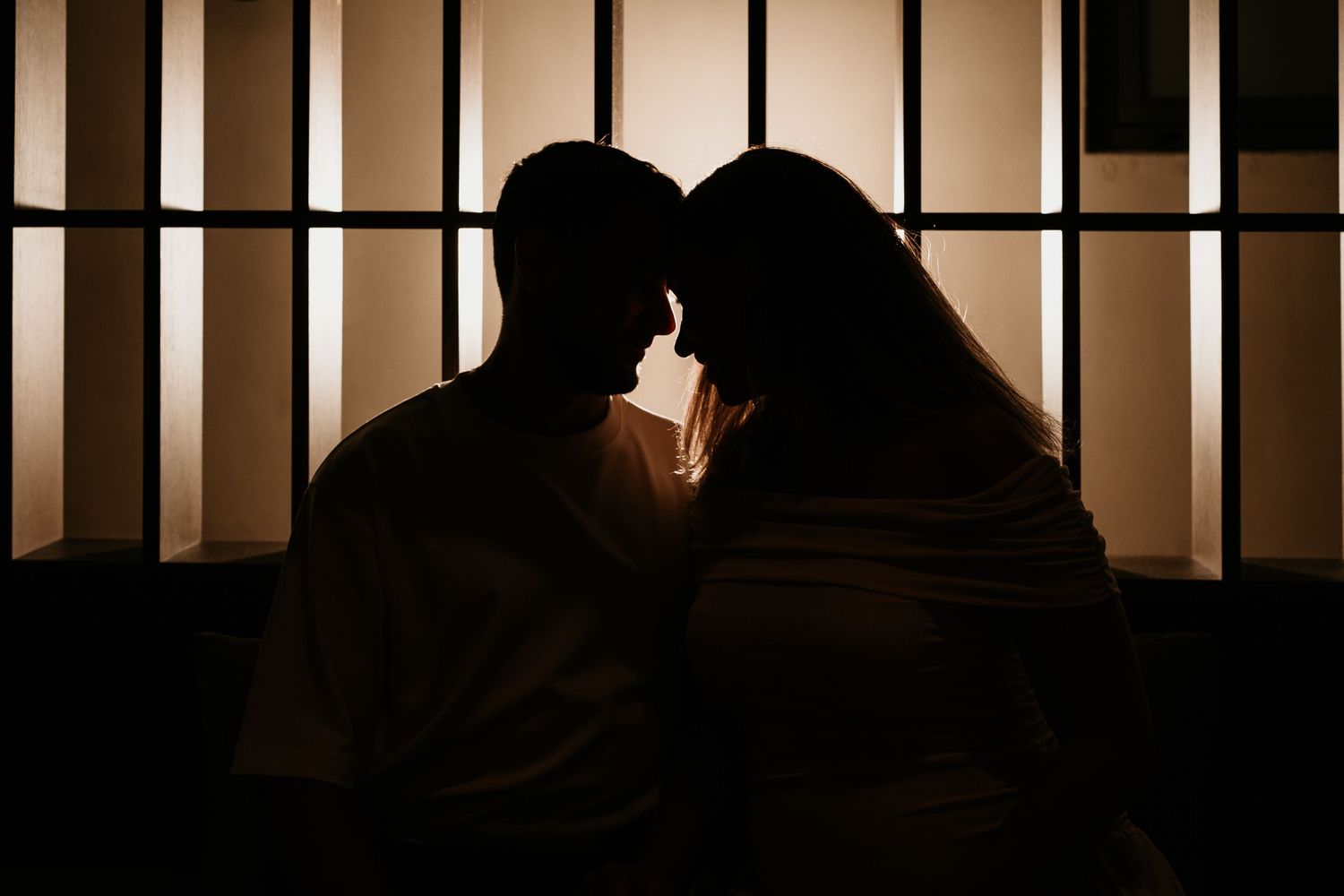 Silhouettes of a couple in a romantic pose against a backlit window with vertical bars.