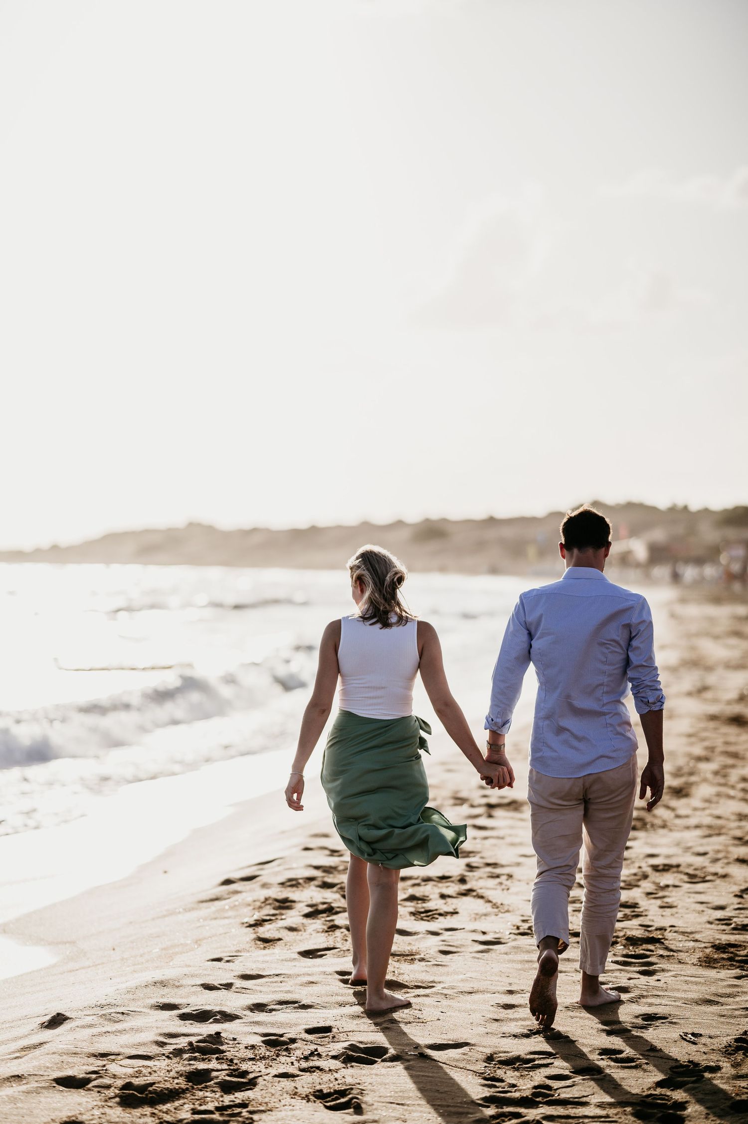 Couple walking hand in hand along a sunset beach with waves crashing nearby.