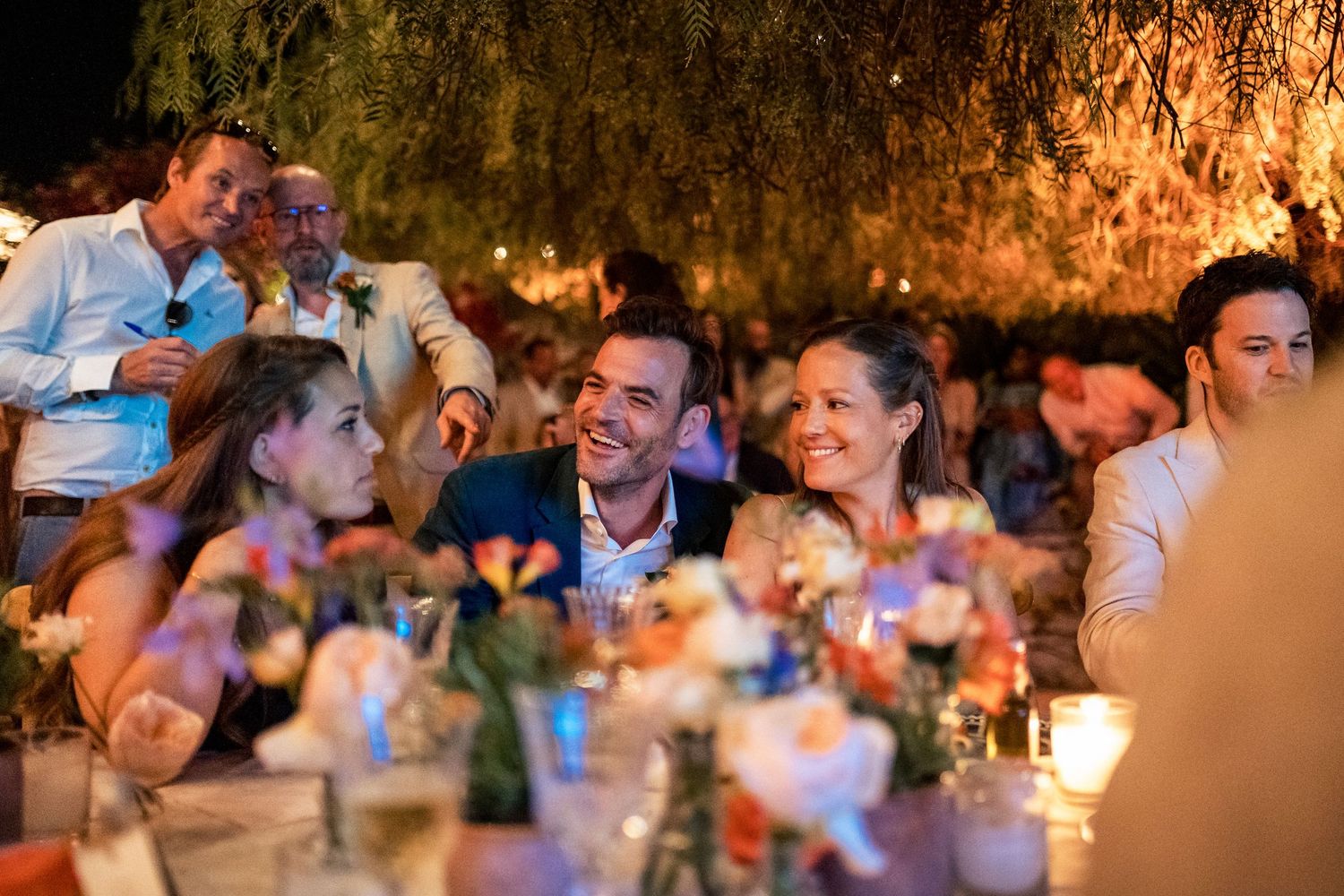 Wedding guests enjoying an outdoor evening reception with warm lighting and natural decor.