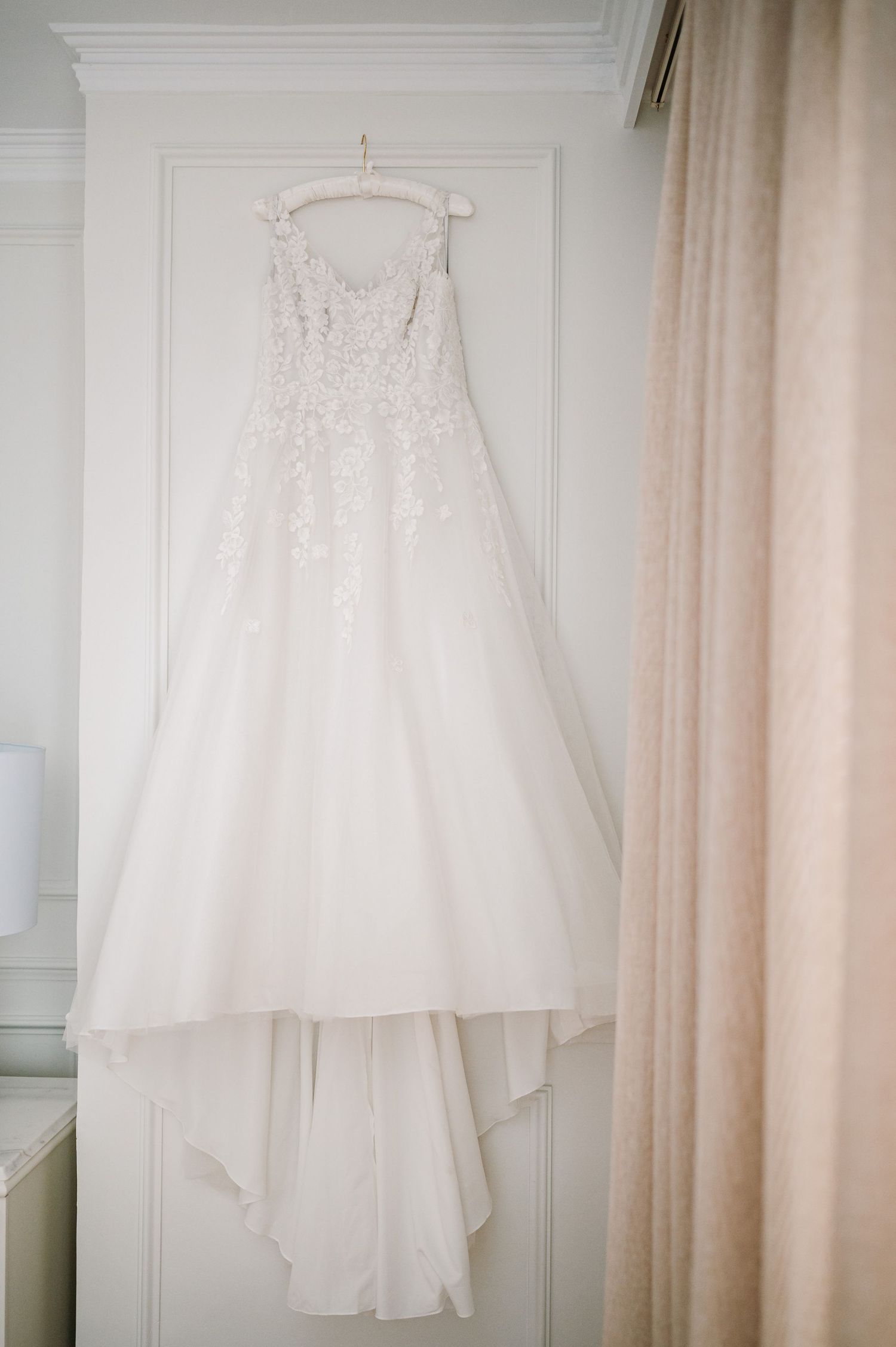 Elegant white wedding dress hanging against soft pink curtains in natural light.