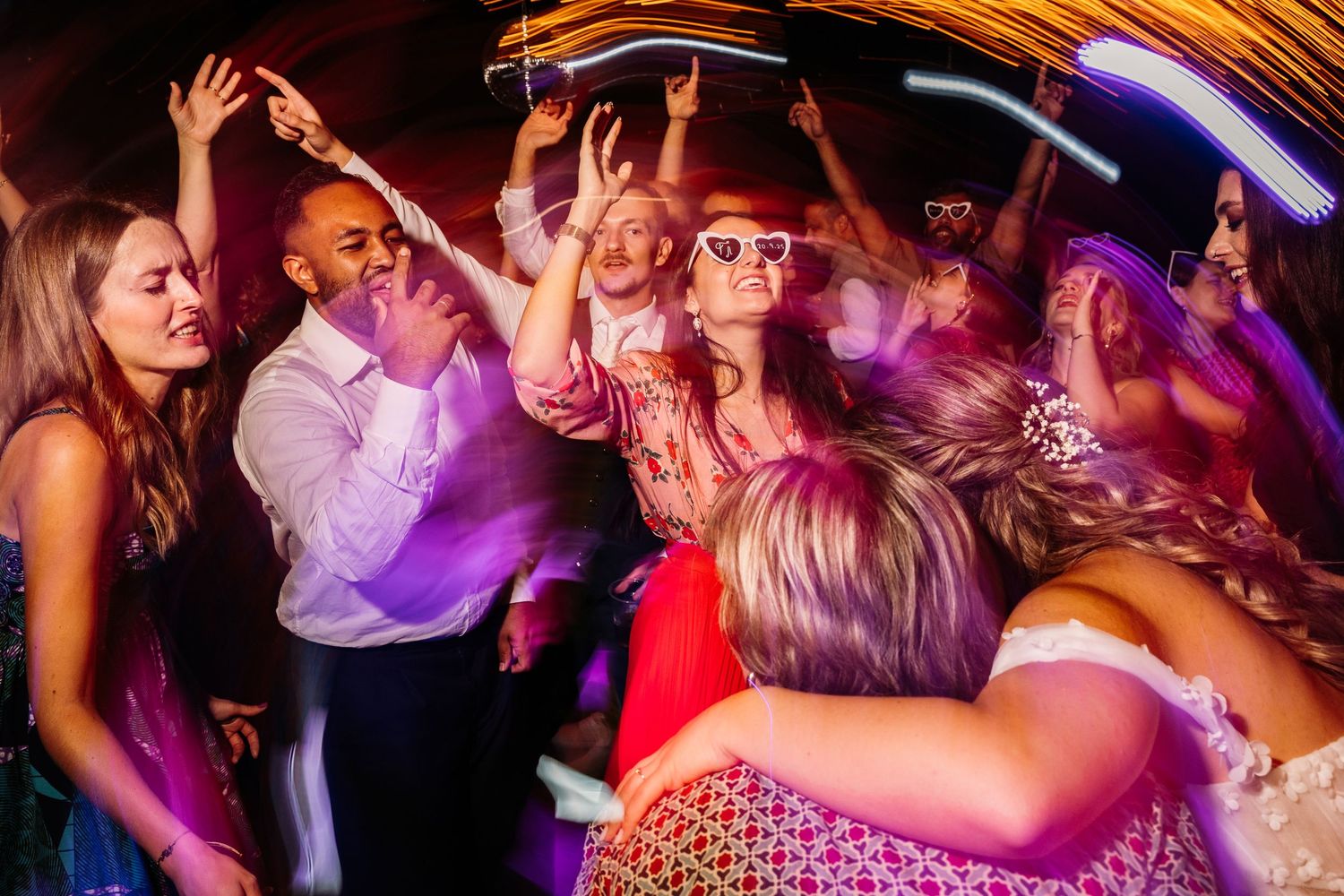 Wedding reception dance floor filled with guests dancing under colorful party lights.